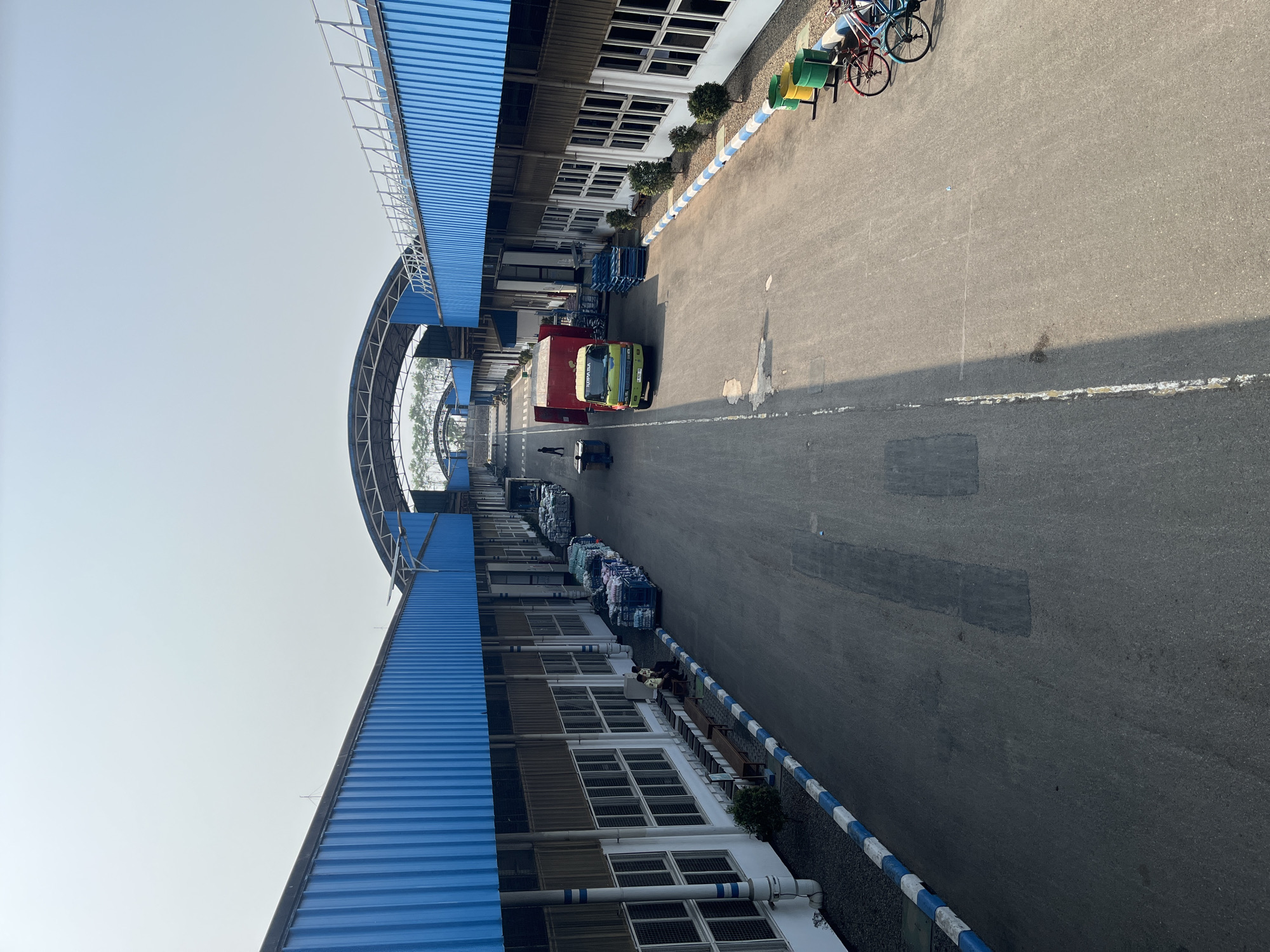 This image depicts an industrial complex, likely a warehouse or distribution center, in Pelang, Indonesia. The scene is dominated by long, low-rise buildings with striking blue corrugated metal roofs and siding. These structures extend along a wide, paved driveway or loading area. Multiple loading docks are visible along the buildings, with some featuring open doors.
On the left side of the driveway, there are rows of stacked goods, some of which appear to be on pallets or in large bags. Benches are positioned along the building's exterior, and two individuals, possibly workers, are seated on one of them. A small green bush is also present.
On the right side of the driveway, there are more stacked goods, including what appear to be large rolls of material and smaller packaged items. Further down, a green and yellow truck with red accents is parked, facing towards the buildings. Next to the truck, a stack of blue pallets is visible. In the distance, more of the blue-roofed buildings stretch out, creating a sense of scale and repetition.
At the far right of the image, three bicycles are parked near some yellow and green cylindrical bins. The ground is paved asphalt, with a blue and white striped border running along the edge of the driveway. The sky is clear and bright, suggesting a sunny day. The overall impression is one of a functional, organized industrial environment.