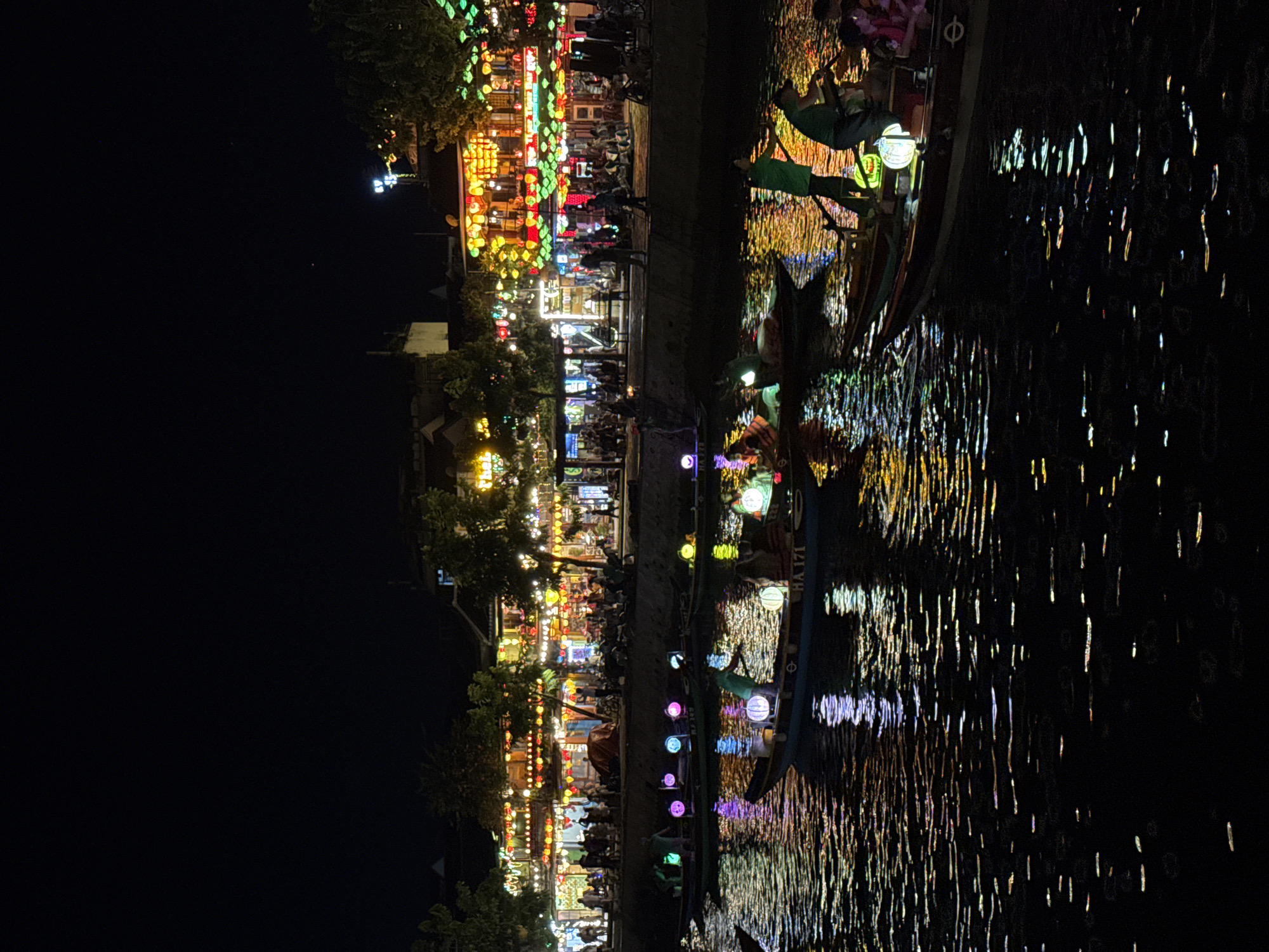 The image depicts a nighttime scene of the Thu Bồn River in Hội An Ward, Vietnam, showcasing the illuminated Old Town. The upper portion of the frame is a dark, clear sky. Below, the river is filled with traditional wooden boats carrying passengers and operators. Each boat is adorned with multiple glowing, spherical lanterns in various colors including pink, purple, blue, green, yellow, and white. Operators, typically standing, propel the boats with long poles. One boat in the mid-ground bears the registration "HA 171". The river's surface reflects the vibrant lights from the boats and the shore, creating elongated, shimmering streaks of color.
Along the riverbank, a promenade is visible, populated by numerous individuals walking, standing, and observing the scene. Behind the promenade, traditional multi-story buildings characteristic of Hội An are extensively illuminated with a dense array of colorful lanterns of various shapes and sizes (predominantly red, yellow, orange, green, and blue). These lanterns are strung across building facades, hung from balconies, and adorn entrances, creating a brightly lit, festive backdrop. Trees are interspersed along the riverbank and among the buildings, partially lit by the lanterns. A bright streetlamp with multiple bulbs is visible on the upper right, contributing to the ambient light. The scene depicts active nighttime tourism and local commerce along the waterway.