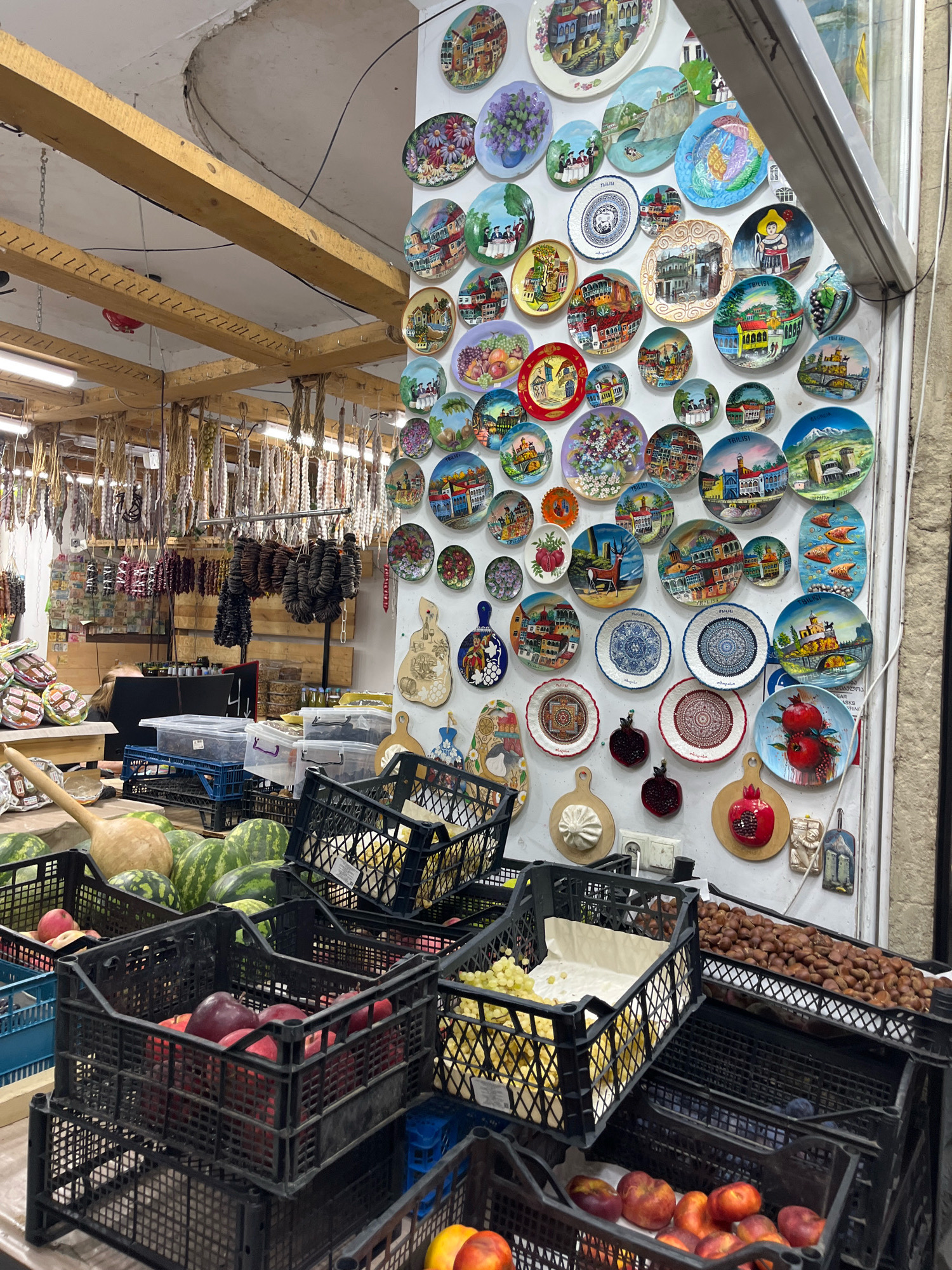 The image depicts a vibrant market stall, likely in Tbilisi, Georgia, given the prominent display of decorative plates featuring Georgian landmarks and traditional motifs. The stall is laden with a variety of goods. In the foreground, several black plastic crates are filled with fresh produce, including watermelons, red apples, and peaches. Behind the fruit, a wooden ladle rests on a pile of watermelons. To the right, more crates contain grapes and a large quantity of nuts, possibly chestnuts.
The wall behind the produce is densely adorned with colorful decorative plates, each depicting different scenes, some identifiable as famous Tbilisi locations like the Narikala Fortress and the sulfur baths, along with artistic representations of fruits, animals, and floral patterns.
Further into the stall, hanging from wooden beams, are numerous strings of dried food items, resembling sausages or jerky, and possibly some dried fruits or vegetables. There are also shelves with packaged goods and other market items in the background. The environment appears to be an indoor market with exposed wooden beams and lighting fixtures overhead. One person, partially visible, is seated at a counter in the background, interacting with the merchandise. The overall impression is of a bustling marketplace selling local crafts and fresh food.