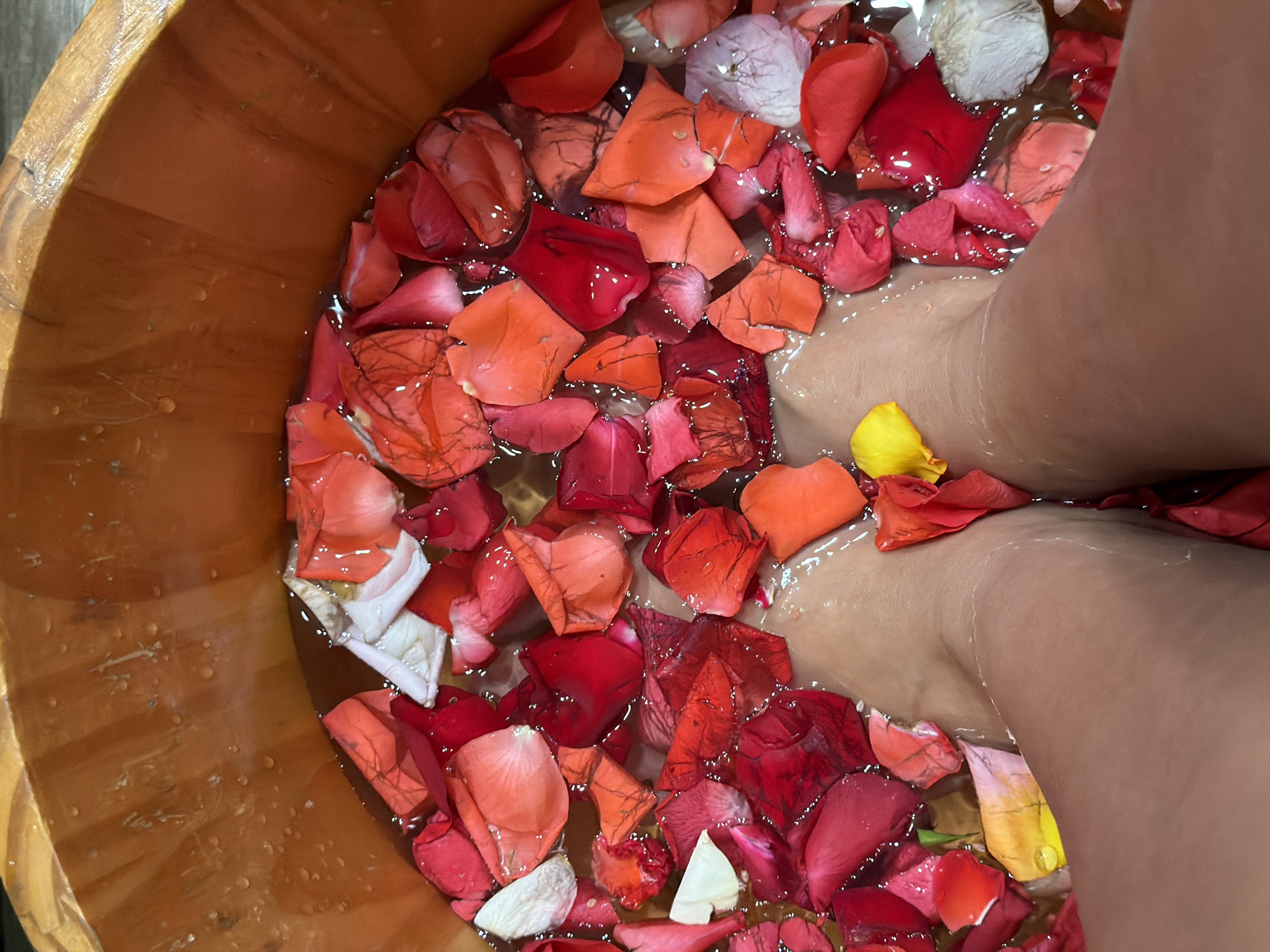 The image depicts a close-up, overhead view of a person's feet submerged in a wooden basin filled with water and numerous rose petals. The rose petals are predominantly red, with accents of pink, orange, and white, and a single yellow petal is visible. The wooden basin has a rough texture and appears to be an outdoor setting, potentially a spa or a private garden area. The water is clear, and the lighting suggests it is daytime. There are no identifiable landmarks or specific details that would indicate the location within Nha Trang Ward, Vietnam. The focus is entirely on the sensory experience of the foot bath.