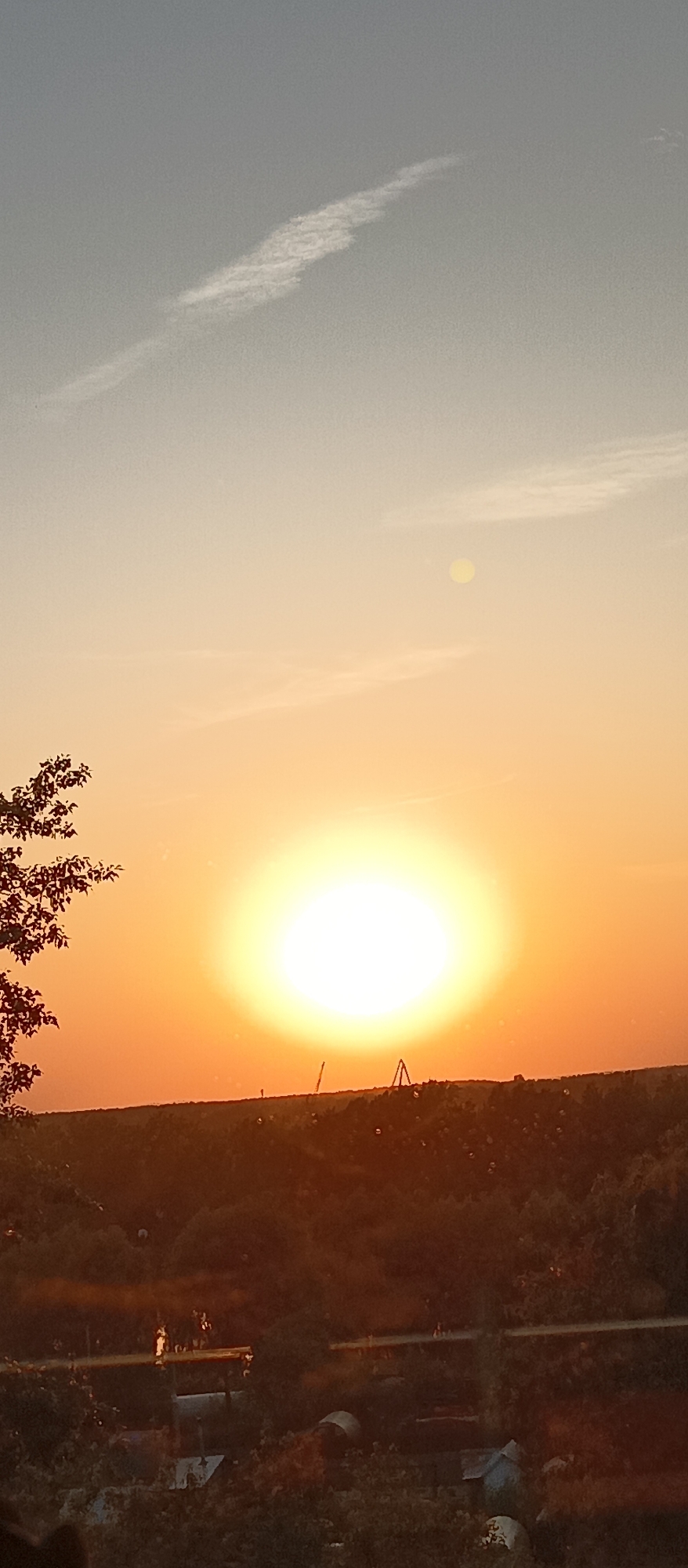 The image shows a landscape scene during sunset. The sun is the dominant feature, appearing as a bright, white-yellow orb in the center, with its glow spreading into the surrounding orange sky. Above the sun, the sky fades into a pale blue-gray color, with a faint white contrail visible in the upper left. Below the sun, a dark, tree-covered land is visible. At the very bottom of the frame, some indistinct, dark objects are partially visible. The location is unknown.