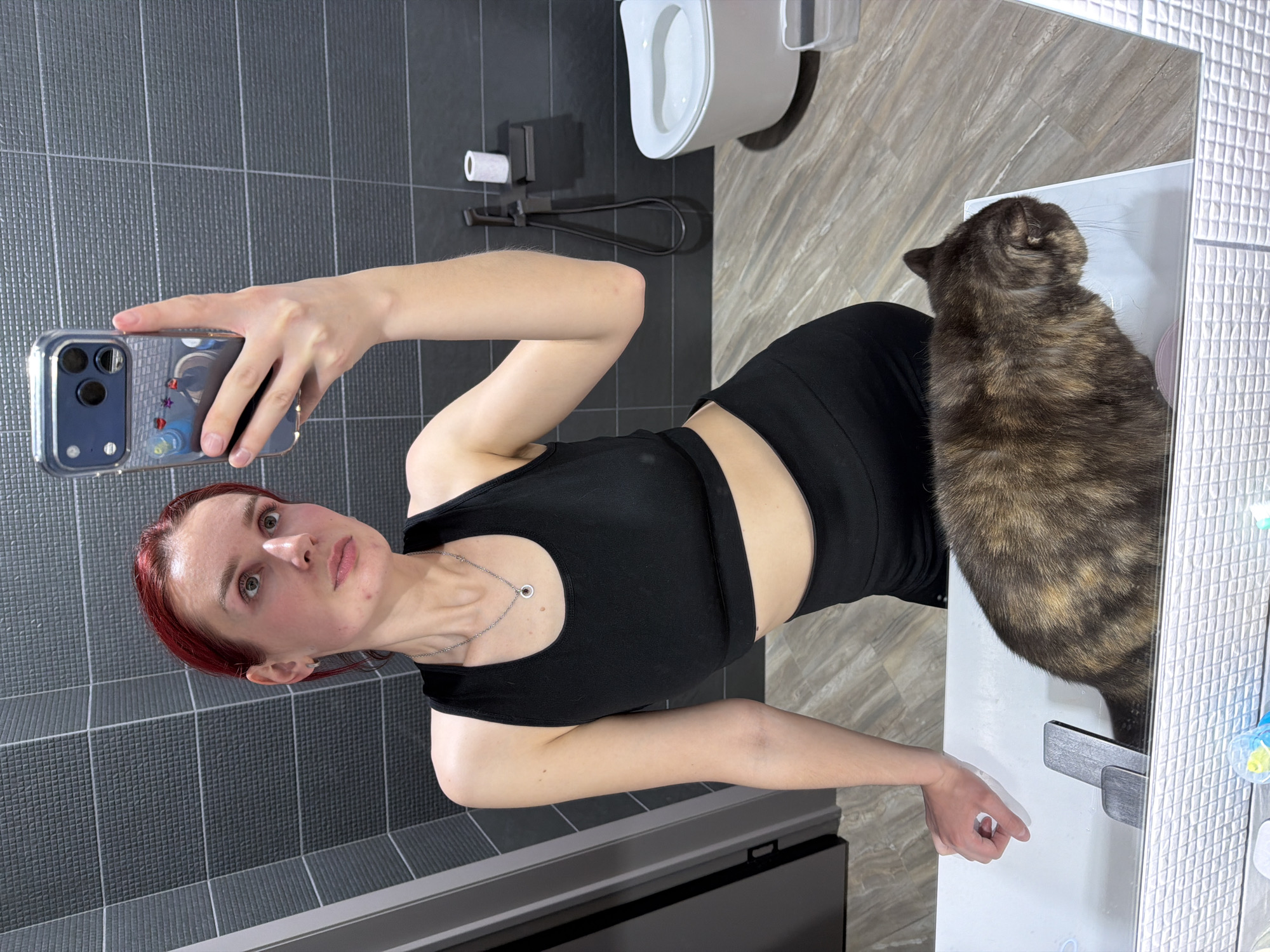 A woman with red hair takes a mirror selfie in a modern bathroom. She is wearing a black athletic top and has a silver necklace with a small dark pendant. Her expression is neutral as she looks into the smartphone she holds horizontally with her right hand. The phone, which appears to be an iPhone, has a clear case with small decorative elements like stars.
To the woman's right, a tortoiseshell cat (brown, black, and tan fur) sits on a white counter or sink, with its back facing the camera.
The bathroom features dark grey, textured rectangular tiles on the wall behind the woman and lighter, marbled brown and beige tiles on the wall behind the cat and on the floor. In the background, mounted on the marbled tiled wall, a white toilet with an open lid is visible. Below the toilet, there is a black toilet paper holder with a roll and a black bidet sprayer.
The scene depicts an indoor setting, likely during the day, with no specific weather visible. No text is discernible in the image. The location context is Chaikovsky, Russia.