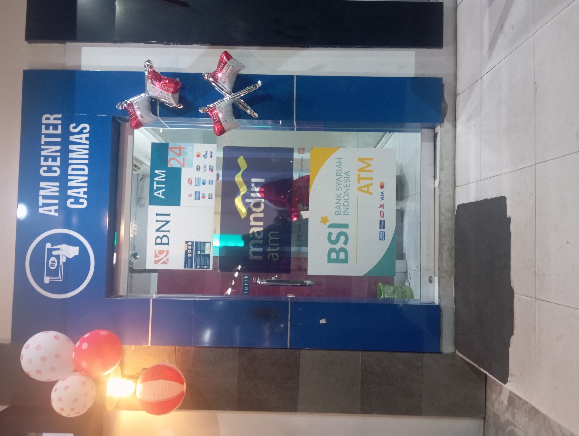 The image shows an ATM center with the name "ATM CENTER CANDIMAS" above the entrance. The entrance is a glass door with various ATM logos and bank names visible, including BNI, Mandiri ATM, and Bank Syariah Indonesia ATM. There are decorative elements like balloons and a paper lantern near the entrance. The floor outside the ATM center is tiled. The location is identified as Purwanegara, Indonesia.