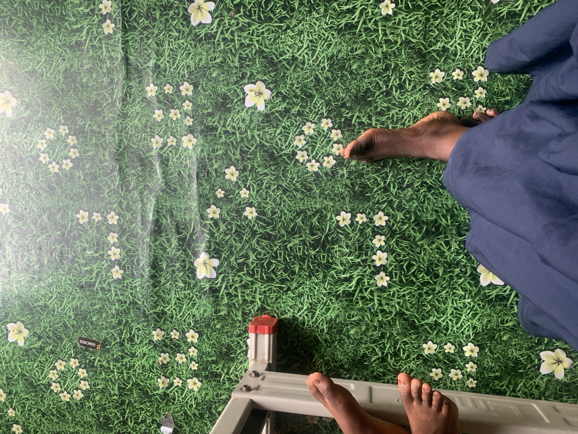 The image is an overhead shot of a person's bare feet standing on a green, grass-like floor covering decorated with scattered white and yellow flowers. A portion of a grey ladder with red accents is visible on the left side of the frame, with the person's left foot positioned near it. The person is wearing dark blue fabric that drapes over their legs. The overall impression is of a playful or whimsical indoor setting, possibly a child's play area or a themed room, as the grass and flowers appear to be a printed surface rather than natural vegetation. There is no discernible indication of the image being located in Jauro Yinu, Nigeria.