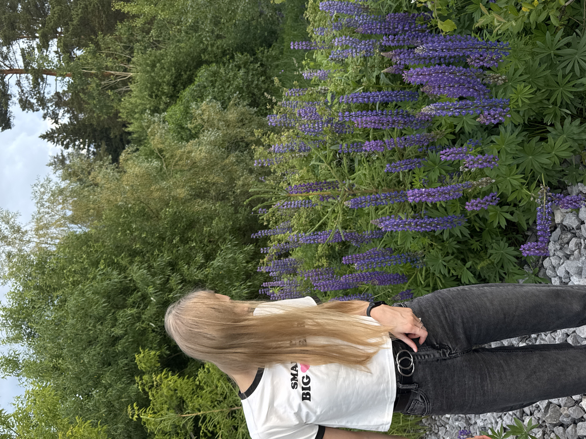 A young Caucasian female with long, blonde hair stands in a natural setting, facing away from the camera and looking towards a field of purple lupine flowers. She is wearing a white crop top with black trim and the words "SMALL BUT BIG" printed on it, along with a pink heart emoji. She also wears dark grey jeans and a black belt with a double-ring buckle. The background consists of lush green trees and foliage. The ground is covered in grey gravel. The scene appears to be a park or natural area on the outskirts of Perm, Russia.