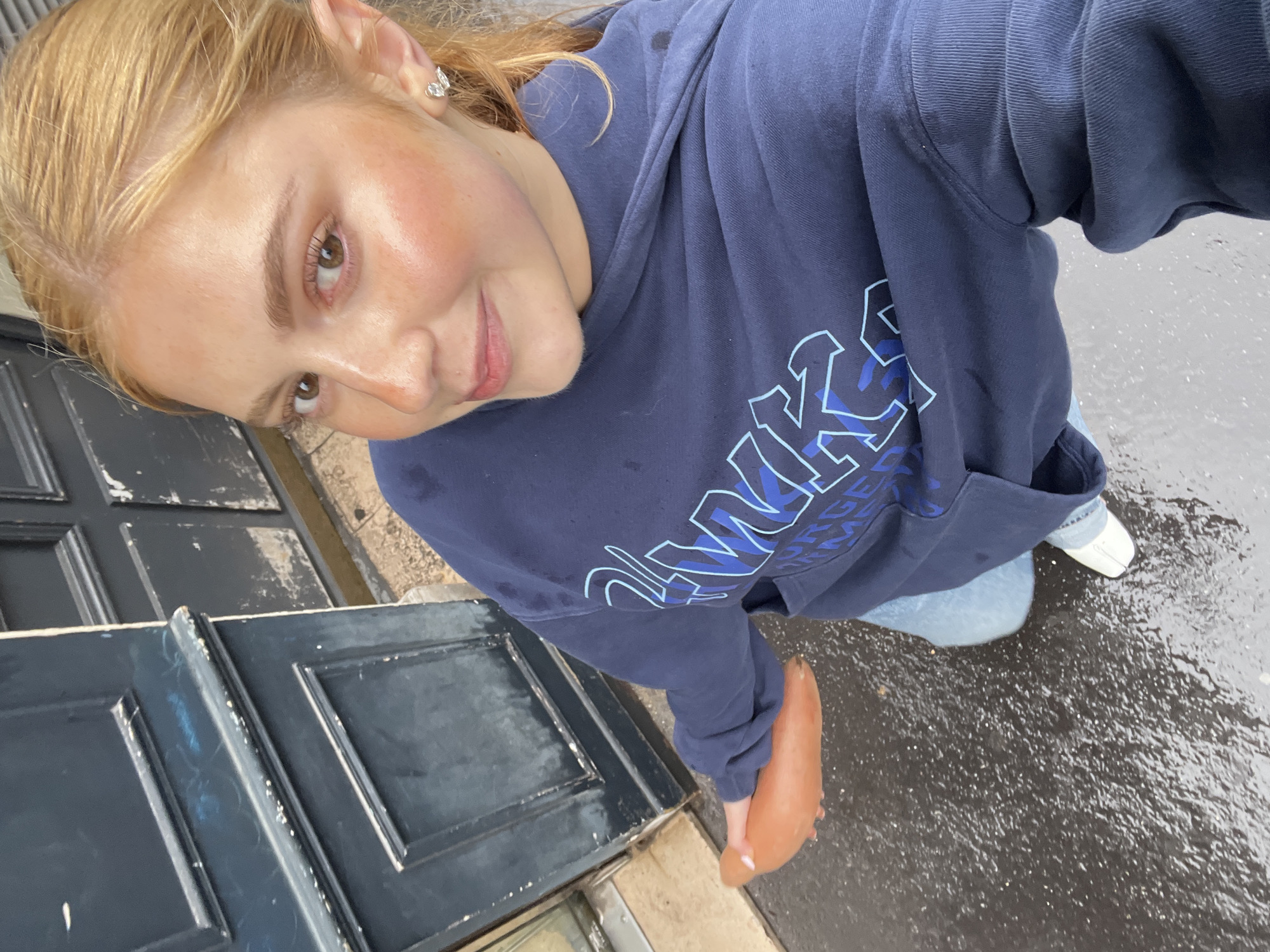 A fair-skinned woman with blonde hair tied back is taking a selfie outdoors. She wears a navy blue hooded sweatshirt with light blue graphics, light blue jeans, and white shoes. She stands on a wet surface, possibly a sidewalk or street, alongside a dark-colored door. The setting appears to be urban. There is insufficient information to determine if the location is Paris, France.