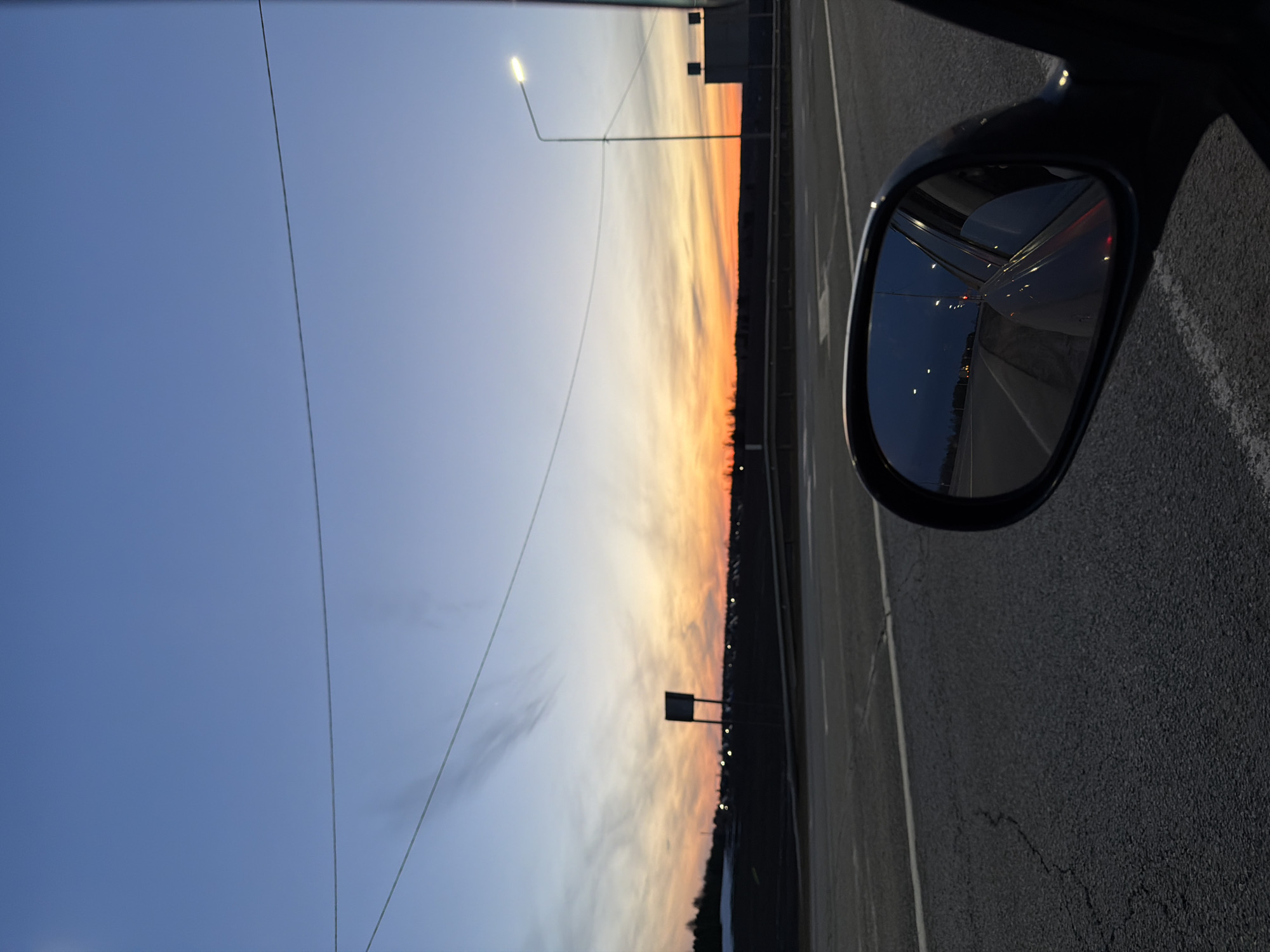 The image is taken from inside a vehicle, potentially a car, while on a bridge or overpass. The right side of the frame features a car's side mirror reflecting a dark highway illuminated by streetlights. To the left, the sky transitions from a light blue at the top to a fiery orange and yellow near the horizon, indicating either a sunrise or sunset. The dark silhouette of trees or a treeline is visible against the horizon. A few utility poles and streetlights punctuate the scene. The image is rotated ninety degrees. Without additional context or geographical landmarks, specifically pinpointing the location within Kuregovo, Russia, is impossible. However, the scene depicts a roadway, likely a highway or main road, possibly on the outskirts of the city, given the presence of a treeline, coupled with an expansive view of the sky and sunset/sunrise.