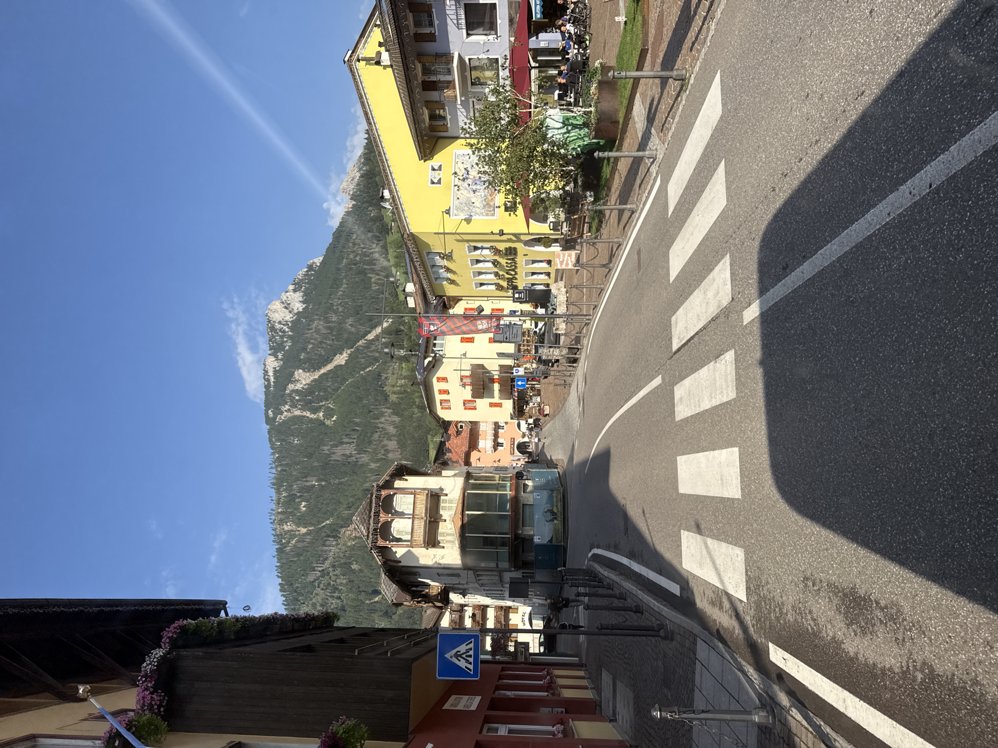 The image shows a street scene in Moena, Italy, likely captured in the morning or late afternoon given the strong shadows. In the foreground, a pedestrian crosswalk is marked on an asphalt road. The street is bordered by buildings on either side. On the left, a dark brown wooden building with flower boxes filled with pink and white flowers is prominent, with a red-painted building adjacent to it. A blue pedestrian crossing sign is visible. On the right, a series of colorful buildings, including a bright yellow one with a mural, line the street, featuring awnings and outdoor seating areas for cafes or restaurants. In the background, a steep, tree-covered mountain rises against a clear blue sky with a faint white contrail. The overall impression is of a charming mountain town bathed in sunlight. There are a few people visible in the distance on the sidewalk near the outdoor seating areas.