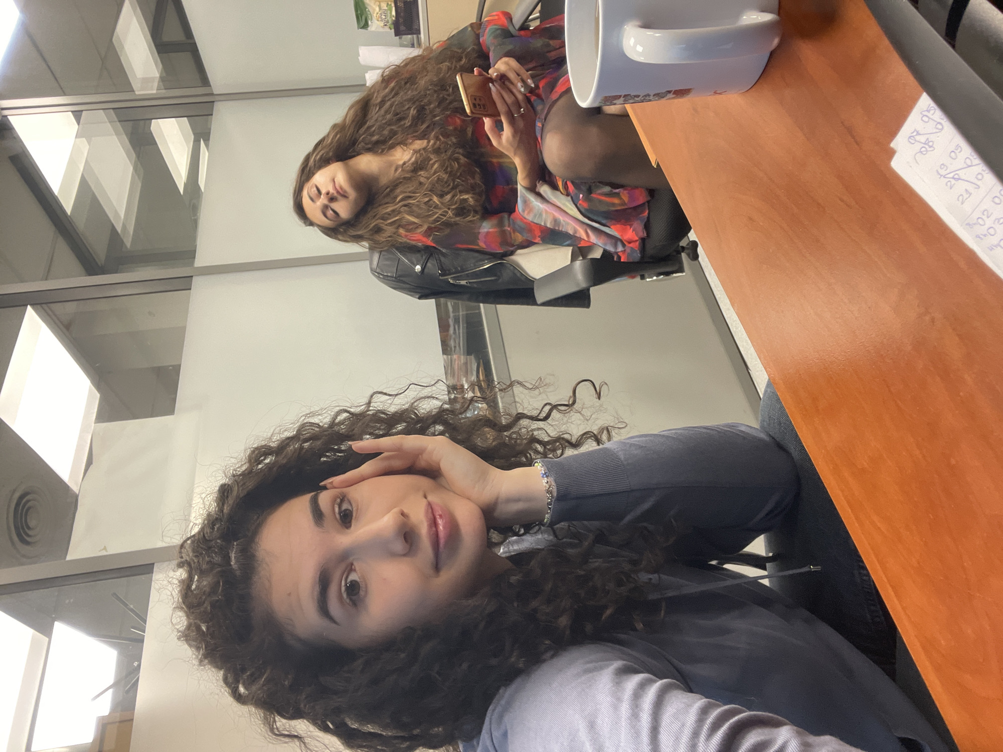 The image portrays two young women in a casual indoor setting, likely an office or commercial building in Sofia, Bulgaria. The photo appears to be a selfie taken from a low angle.
In the foreground, a woman with voluminous dark, curly hair looks directly at the camera with a relaxed expression and a slight smile. Her left hand is raised to her temple, with fingers resting on her forehead. She is wearing a dark blue or grey long-sleeved top, possibly a hoodie, and a light-colored, multi-beaded bracelet on her left wrist.
In the background, a second woman is seated in a black office chair, reclining. She has long, wavy brown hair and is dressed in a colorful, patterned long-sleeved top or dress, layered with a black leather jacket and dark tights. She holds a gold or tan smartphone in her hands, her gaze directed towards it with a serene expression, eyes partially closed.
The setting features modern interior design with white walls and prominent glass partitions or windows, reflecting internal structures and ambient light, suggesting a daytime environment. To the right, a reddish-brown wooden desk holds a white ceramic mug with a handle. A small piece of white paper on the desk displays handwritten numbers such as "07:05", "19:05", "20:05", and "21:05," possibly indicating a schedule or timestamps. The overall activity suggests a moment of relaxation or a break between the two women.