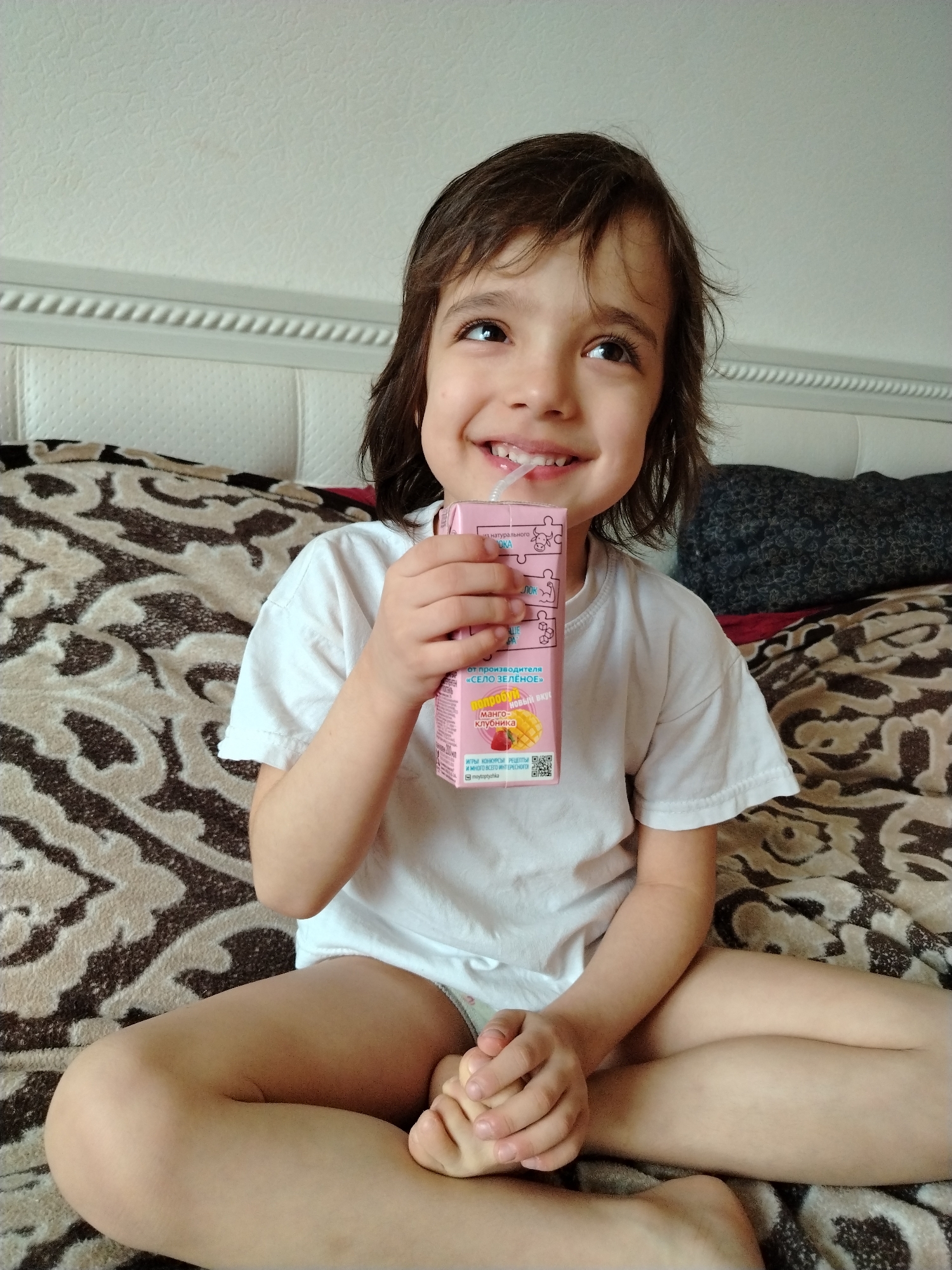 A young child, likely a girl, with shoulder-length dark hair, is captured sitting cross-legged on a bed in Noviy, Russia. She is wearing a white short-sleeved t-shirt and light-colored underwear or shorts. The child holds a pink juice box to her mouth with a straw, appearing to be drinking from it. A wide, happy smile is on her face as she looks upwards and slightly to the right, suggesting a moment of joy, curiosity, or engagement with something out of frame.
The setting is indoors, presumably a bedroom, during the daytime, indicated by the bright, natural-looking light. The bed features a white upholstered headboard with a textured design and decorative molding along its top edge. The child sits on a patterned blanket or comforter, which displays an intricate design in shades of brown, beige, and black.
The pink juice box is a notable object. Visible text on the box, as identified by OCR, includes: "натурального" (natural), "от производителя" (from the manufacturer), and the brand name "«СЕЛО ЗЕЛЕНОЕ»" (Selo Zelenoye, meaning Green Village). The flavor is specified as "Манго-клубника" (Mango-Strawberry). Further text encourages consumers to "попробуй" (try) and mentions "ИГРЫ КОНКУРСЫ РЕЦЕПТЫ И МНОГО ВСЕГО ИНТЕРЕСНОГО" (Games, Contests, Recipes, and much more interesting). A social media handle "moytoptychka" and a QR code are also present. The juice box itself has puzzle-piece-like graphics on its side. The child's expression conveys contentment and playful innocence.