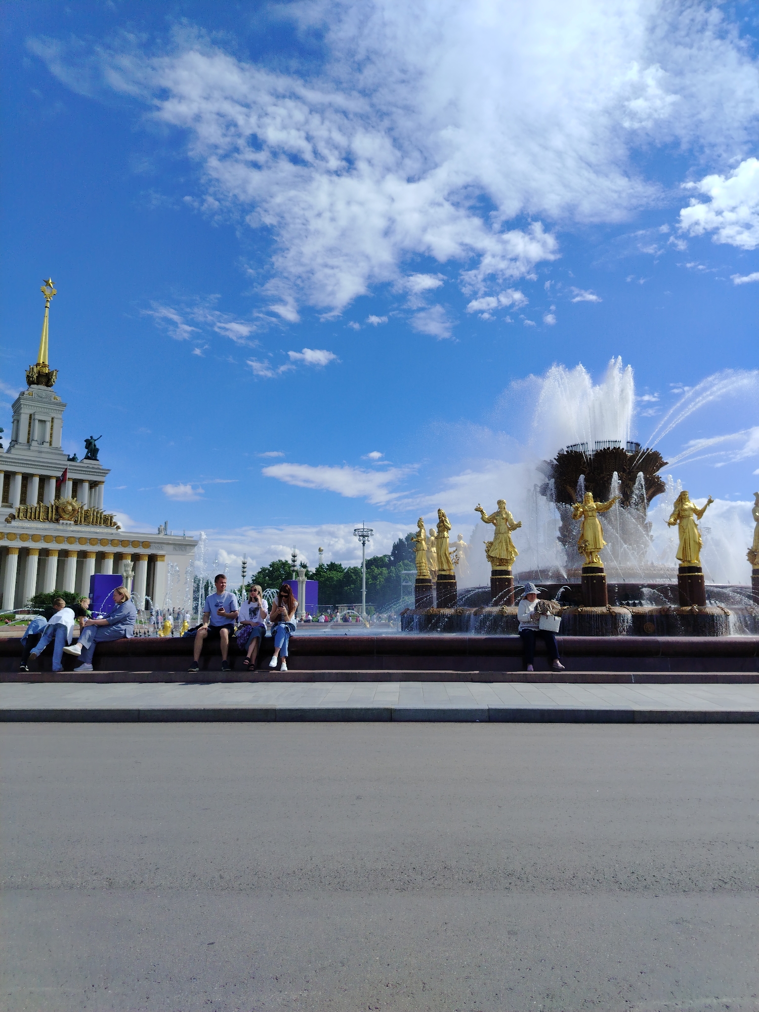 This media file depicts a sunny day scene at a public fountain, likely in Moscow, Russia, given the architectural style and the presence of the "Friendship of Nations" fountain. The fountain is a prominent feature, adorned with numerous golden statues of women, each holding a sheaf of wheat, symbolizing agricultural abundance. Water jets erupt from the center and cascade down the tiers of the fountain.
Several people are casually gathered around the fountain. On the left side, a group of individuals is seated on the fountain's edge, some engaged in conversation, others looking at their phones. Further to the right, more people are also seated, enjoying the view and the atmosphere. One person in the foreground is holding a basket.
In the background, a grand, ornate building with classical architecture stands tall, topped with a golden spire and a star, a common motif in Soviet-era structures. The sky is a vibrant blue, dotted with fluffy white clouds, suggesting fair weather and a pleasant time of day, likely mid-morning to afternoon. The overall mood is relaxed and leisurely, with people enjoying a public space on a beautiful day. The scene evokes a sense of public gathering and appreciation for art and architecture.