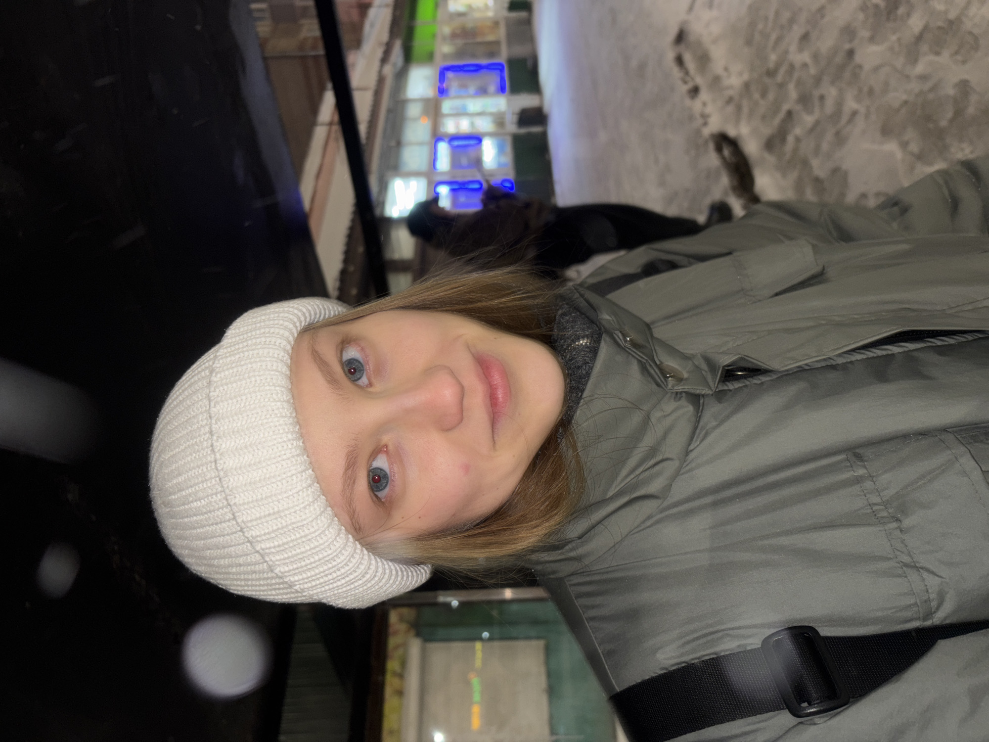 A person with blue eyes, fair skin, and light brown hair is wearing a white knit beanie and a gray hooded jacket with a black strap. The person is facing the camera with a slight smile. The background includes a snowy sidewalk and buildings with illuminated windows, possibly stores or shops. The location appears to be in a city. Based on the available visual information, it is not possible to confirm that the exact location is Perm, Russia, without additional context or data. More information is needed for confirmation.