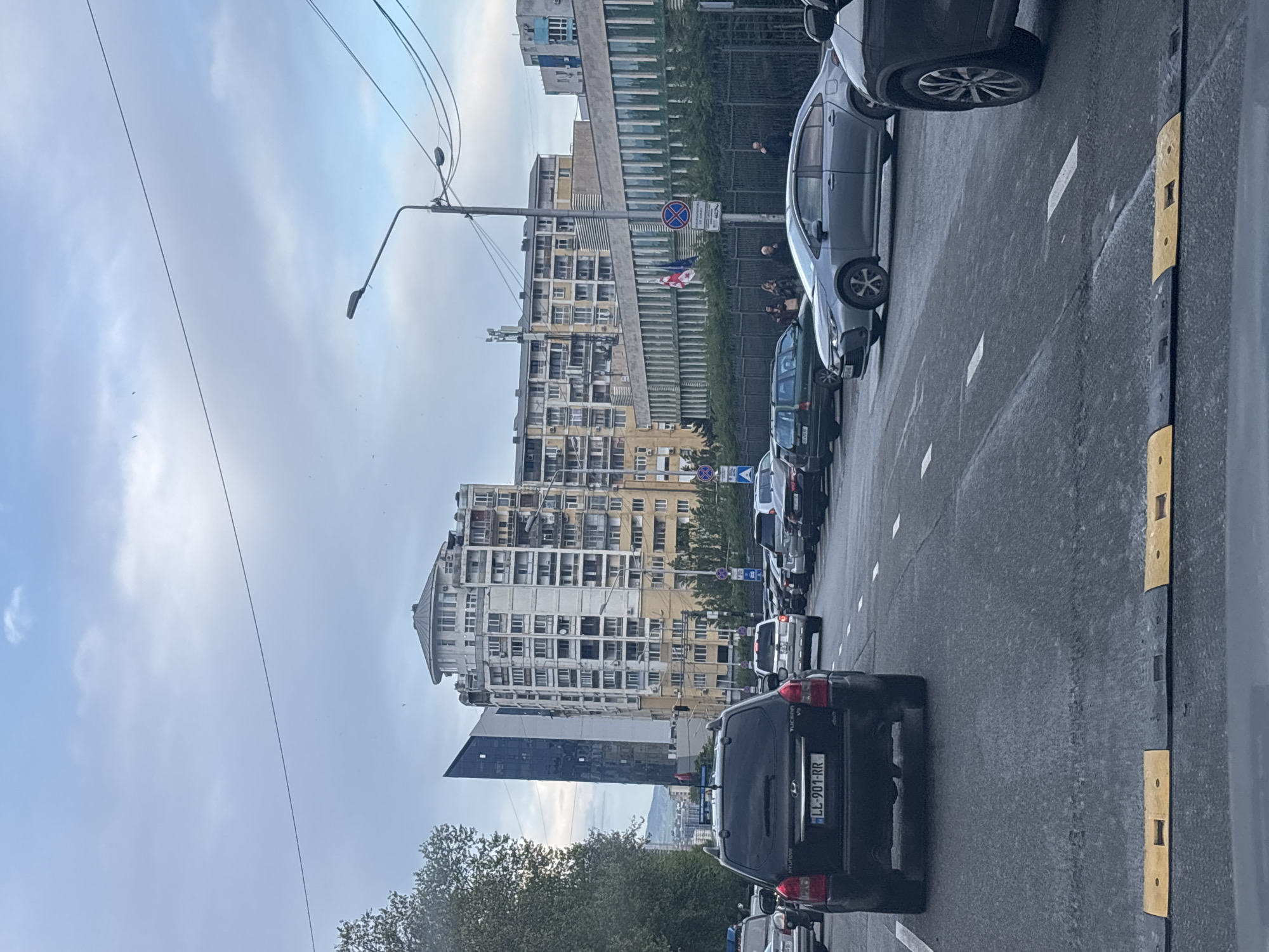 The image shows a street scene in Tbilisi, Georgia, characterized by a traffic jam on a multi-lane road. The foreground features several cars lined up, with a black Hyundai Tucson prominently displayed. Roadside features include lane markings and speed bump dividers. In the background, distinctive architecture is visible, including a tall, light-colored building with numerous windows and a rounded top. A modern glass-paneled building also stands in the background, reflecting the sky. Streetlights and utility lines are noticeable above the traffic. A Georgian flag can be seen at the building, marking its location as Georgia. People are seen gathered at the building, possibly observing the street scene. The sky is overcast with clouds.
