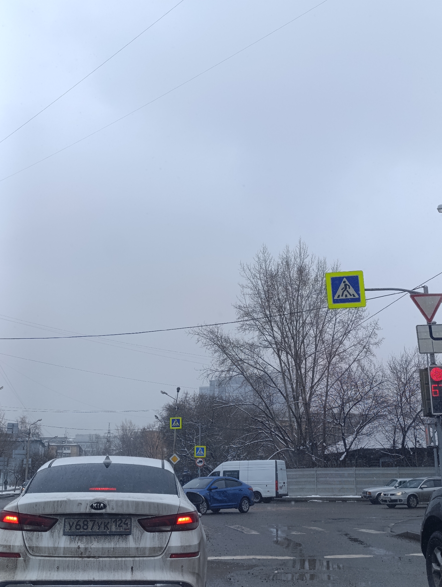 This image depicts a winter street scene at an intersection in Krasnoyarsk, Russia, under a heavily overcast sky. The road surface appears wet and slushy, and snow is visible on the ground and covering the bare branches of deciduous trees, indicating cold weather conditions.
In the foreground, the rear of a white Kia sedan, covered in road spray and dirt, is prominent. Its license plate, "Y687YK 124 RUS", confirms the location, with '124' being the region code for Krasnoyarsk Krai. Further down the street, a blue car is visible with noticeable damage to its front end, suggesting it has been involved in a collision. Behind the blue car, a white cargo van is stopped, and other vehicles can be seen in the distance.
The intersection is equipped with various traffic signs and signals. A large yellow square sign with a black pedestrian symbol is mounted above the road. To its right, a red triangular "Give Way" sign is visible, along with a red traffic light displaying the number "6," likely indicating seconds remaining before it changes. Other signs, including a diamond-shaped priority road sign, are also present. Power lines crisscross the dull sky, and urban buildings are visible in the background, contributing to the city environment. The scene captures a typical, somewhat somber winter day in an urban setting, marked by the aftermath of a minor traffic incident.