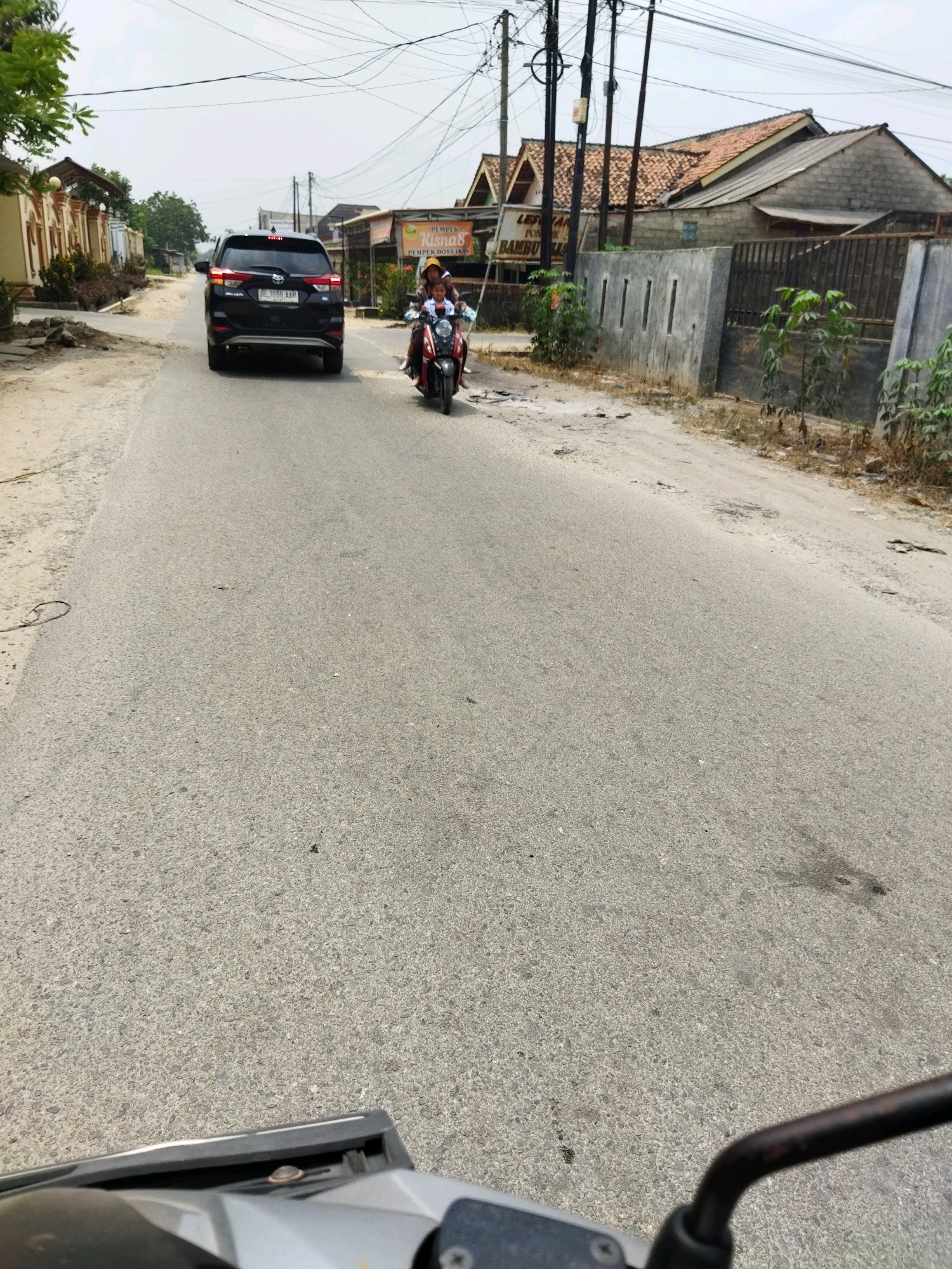 A black SUV is driving down a paved road in Banjar Agung, Indonesia. A motorcycle with two occupants is also on the road, following the SUV. The road is lined with houses and businesses, and there are power lines overhead. The sky is overcast.