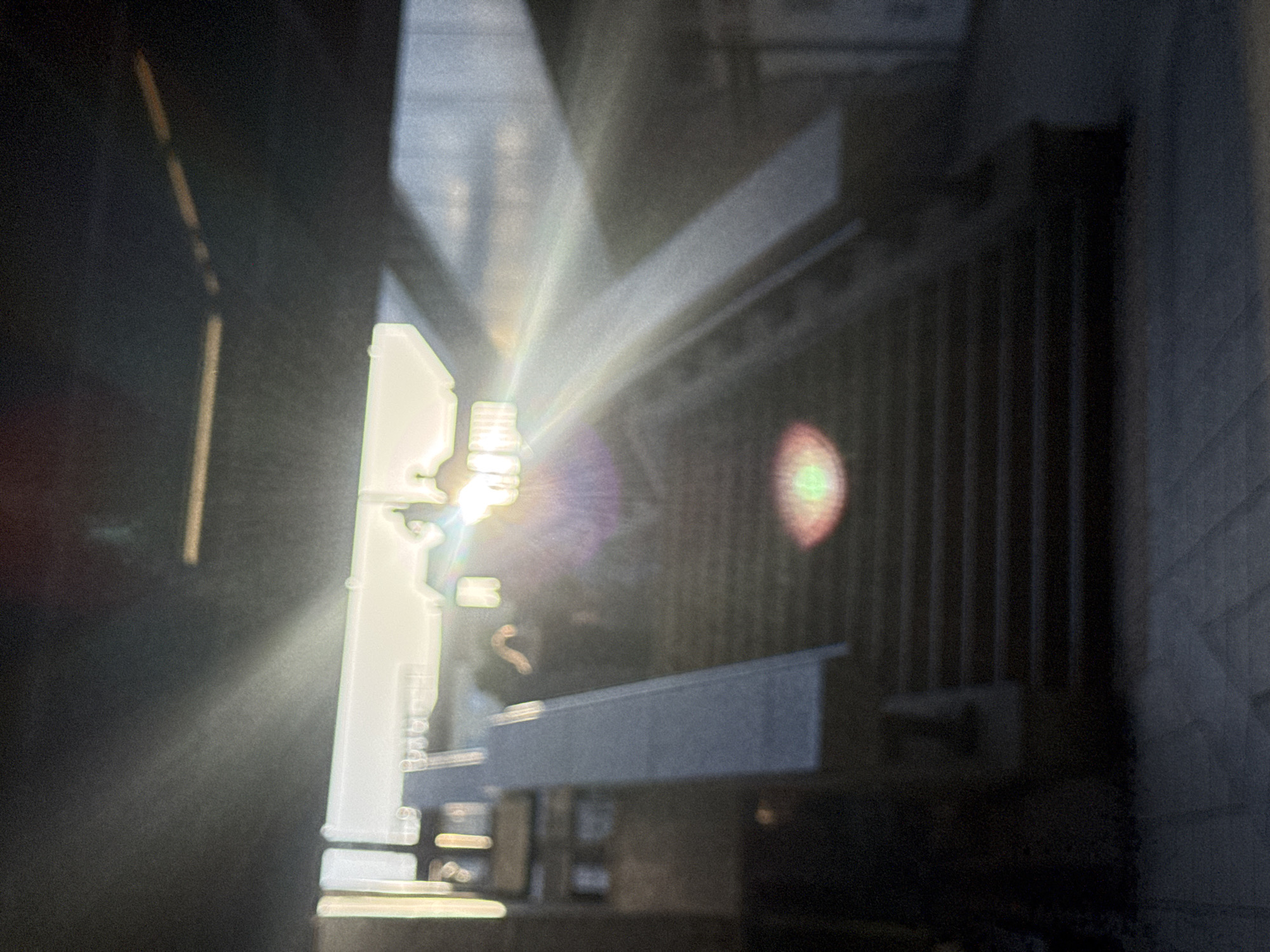 The image shows a blurred perspective view within an urban environment. Strong sunlight obscures the details of the scene. It is difficult to definitively identify the exact location as Sofia, Bulgaria, due to the lack of clear landmarks. The scene includes building elements, possible metallic structures or scaffolding, and a series of vertical bars. A hazy, reddish-green orb appears as lens flare. The overall ambiance is unclear due to blurring and high-contrast lighting. No people are definitively visible.