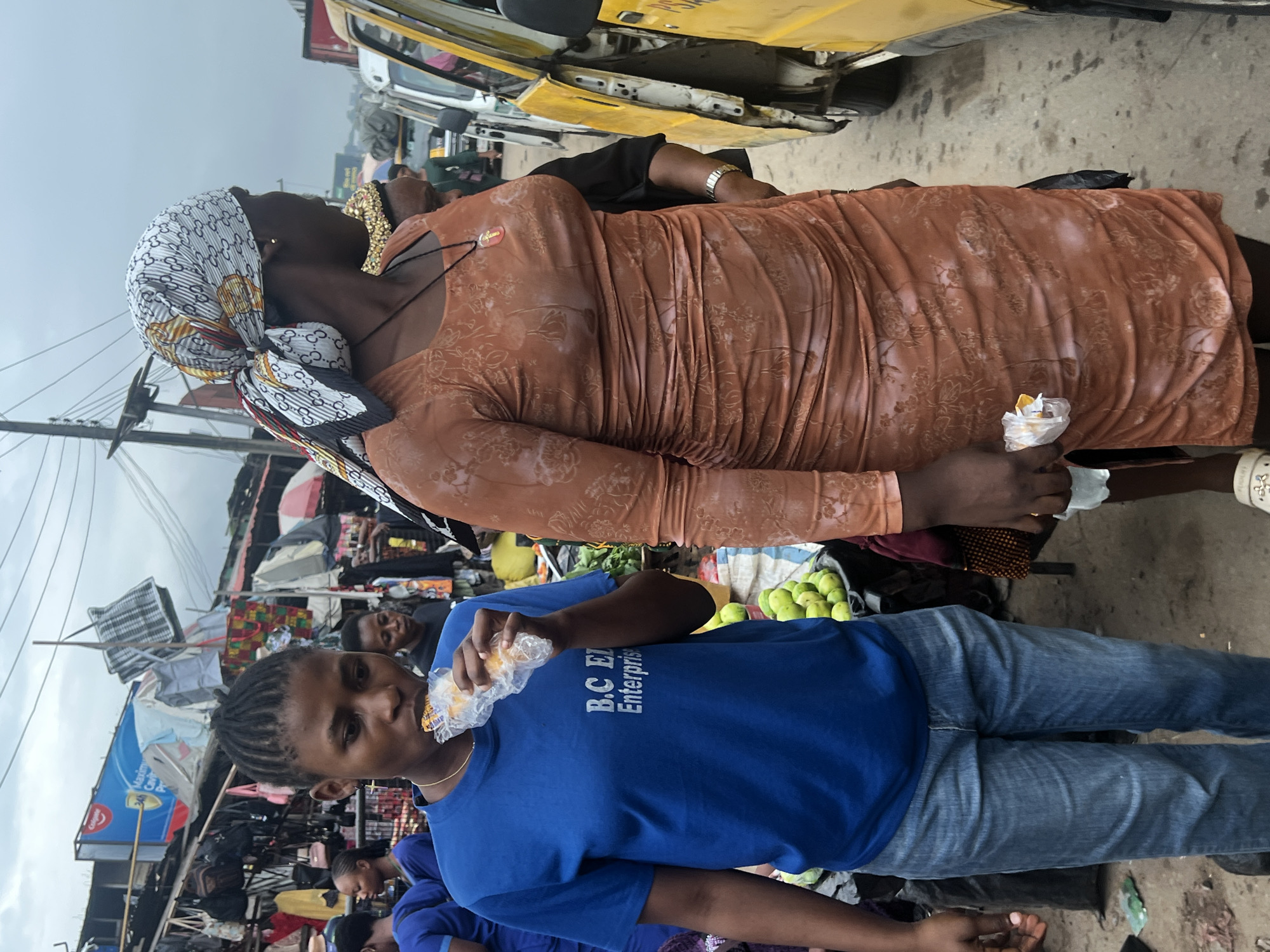 Two women stand on a paved surface in what appears to be a bustling market in Nkpor, Nigeria, under an overcast sky. The woman on the left, wearing a blue t-shirt with "B.C. Enterprise" printed in white and blue jeans, is eating a wrapped pastry. She has braided hair and is facing left. The woman on the right is wearing a patterned orange dress with ruched detailing and a headscarf. She is holding a wrapped pastry in her right hand and is looking to her right, away from the camera. Behind them, the market scene unfolds with stalls displaying various goods, including produce like green fruits in baskets. Vehicles, including a yellow bus, are visible in the background, suggesting a busy urban environment. The ground is dusty and there are scattered items, indicative of a lively marketplace.