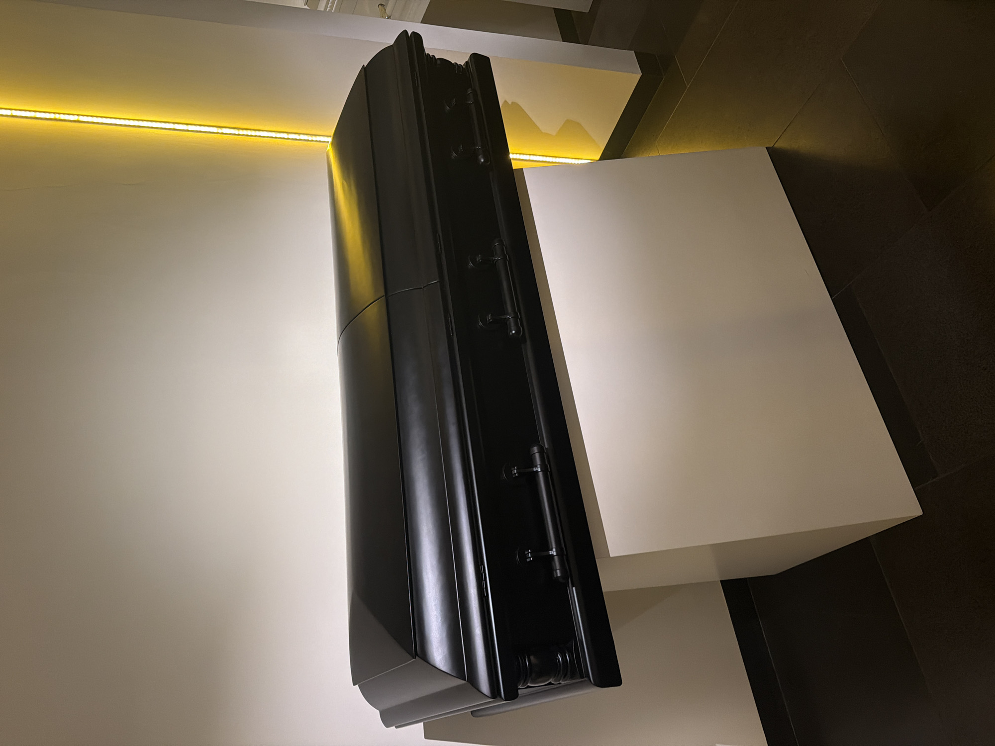 The image shows a black, vertically-oriented cabinet mounted on a white platform. The cabinet is positioned against a white wall with a linear yellow light fixture near the top. The platform is adjacent to a dark-tiled floor. The location is unknown.