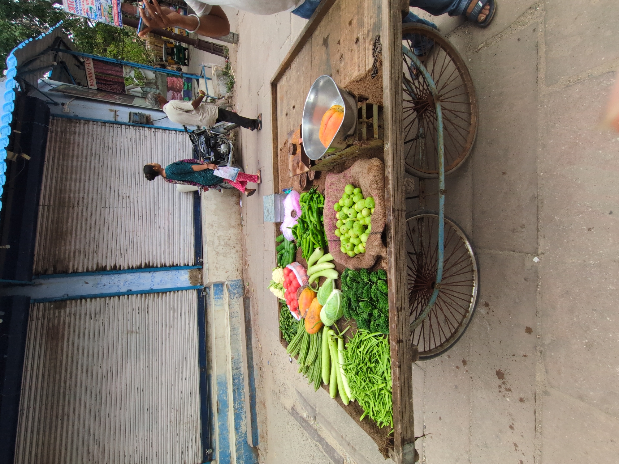 The image depicts a street-level view of a vegetable cart in Jodhpur, India. The cart, laden with an assortment of fresh produce including beans, gourds, tomatoes, cauliflower, cucumbers, and what appear to be papayas, is positioned on a paved street. In the background, a row of shops with closed metal shutters lines the street. Two individuals are walking away from the shops. One is a woman wearing a green and pink patterned outfit, carrying a white plastic bag. The other is an older man in a light-colored shirt and dark pants. A motorcycle is parked near them. The overall scene suggests a typical market setting in a bustling Indian city.