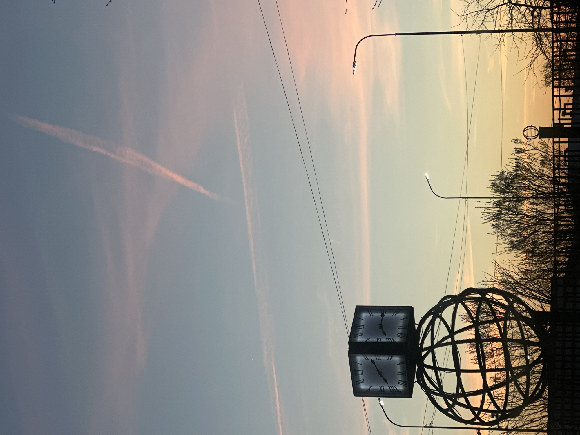 The image shows a view of the sky at sunset in Chaikovsky, Russia. There are pink and orange clouds with contrails from airplanes visible in the sky. In the foreground is a silhouette of a clock and a globe sculpture, along with bare tree branches and utility lines. Streetlights are also visible in the background.