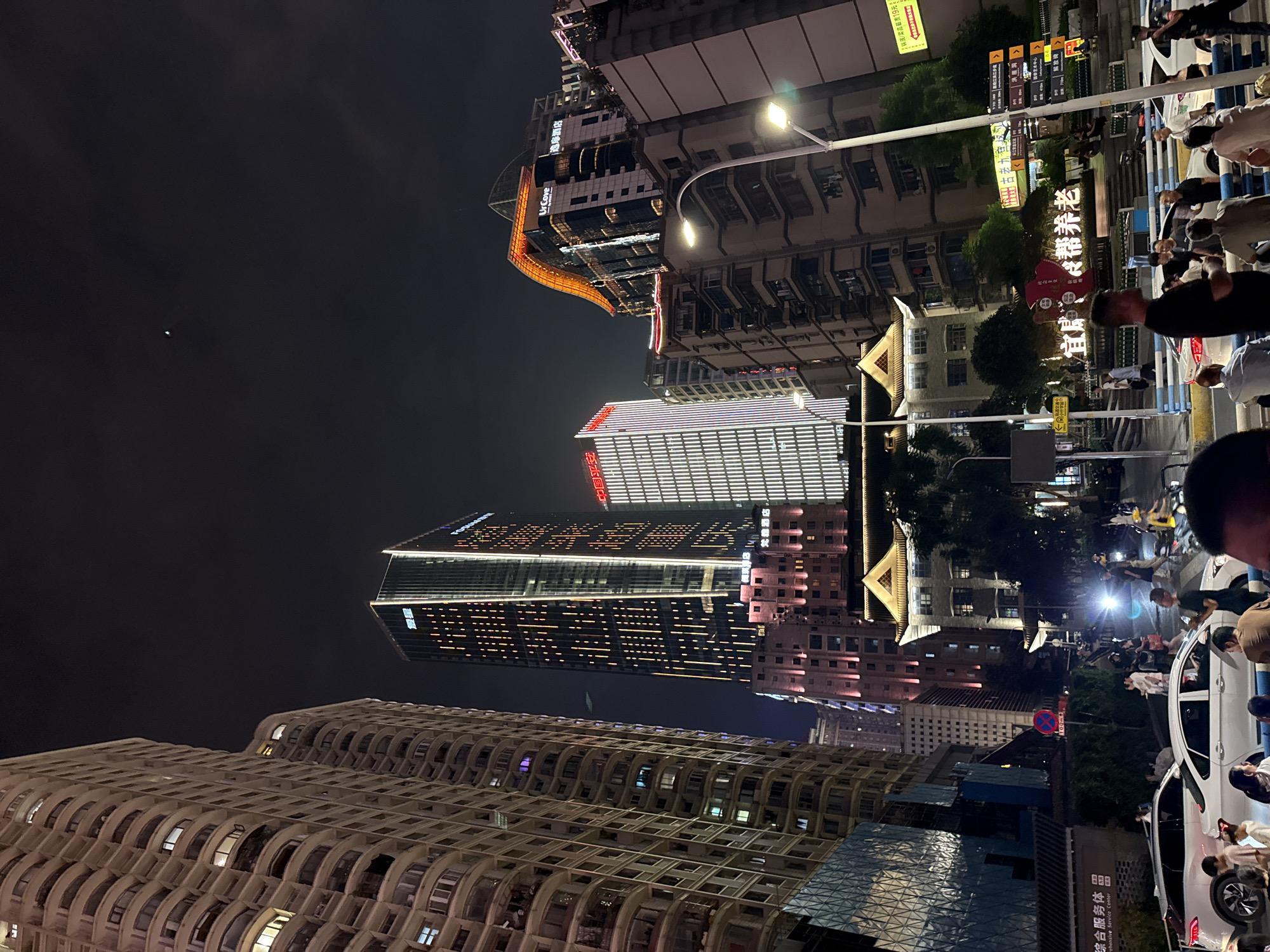 This is a nighttime, low-angle shot of a busy city street in Yuzhong District, China, featuring a mix of modern high-rise buildings and older structures. A tall, slender skyscraper with vertical yellow and white lights dominates the center-left of the frame, displaying Chinese characters at its peak. To its right, another modern building has a distinctive, curved, orange-lit roof. Several other multi-story residential and commercial buildings, illuminated by a variety of lights, fill the background. In the foreground, a street scene unfolds with several white cars, one of which has its headlights on. Pedestrians are visible on the sidewalks and crossing the street, some of whom appear to be gathering or conversing. Streetlights and illuminated signs contribute to the urban glow. The overall atmosphere is one of urban activity at night.