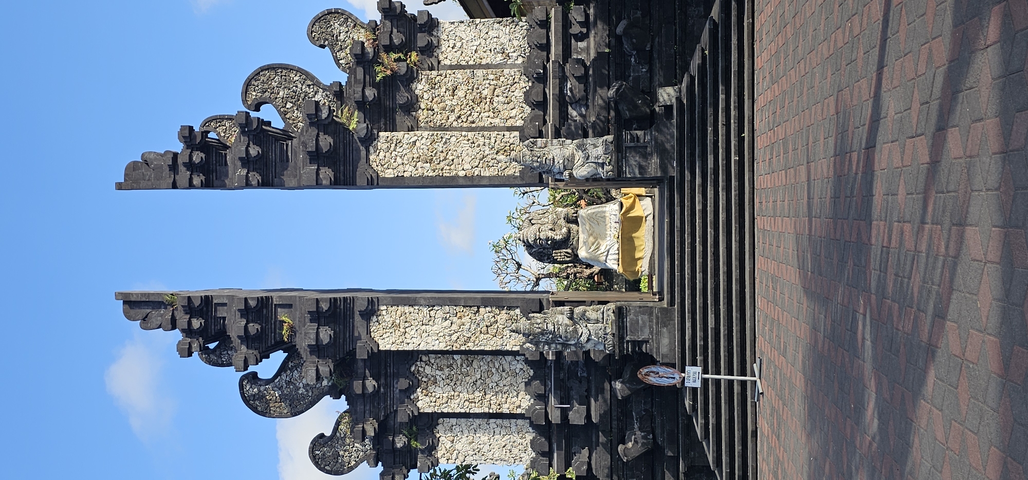 The image depicts a traditional Balinese gate, known as a Candi Bentar, likely located in Denpasar, Indonesia. The gate features black stone carvings with stacked patterns, interspersed with sections of white stone. A stone statue draped in a yellow sash stands at the gate entrance. The gate is flanked by a sidewalk paved with alternating red and gray bricks. A sign with Indonesian text is visible near the base of the gate. The sky above is blue with scattered white clouds.