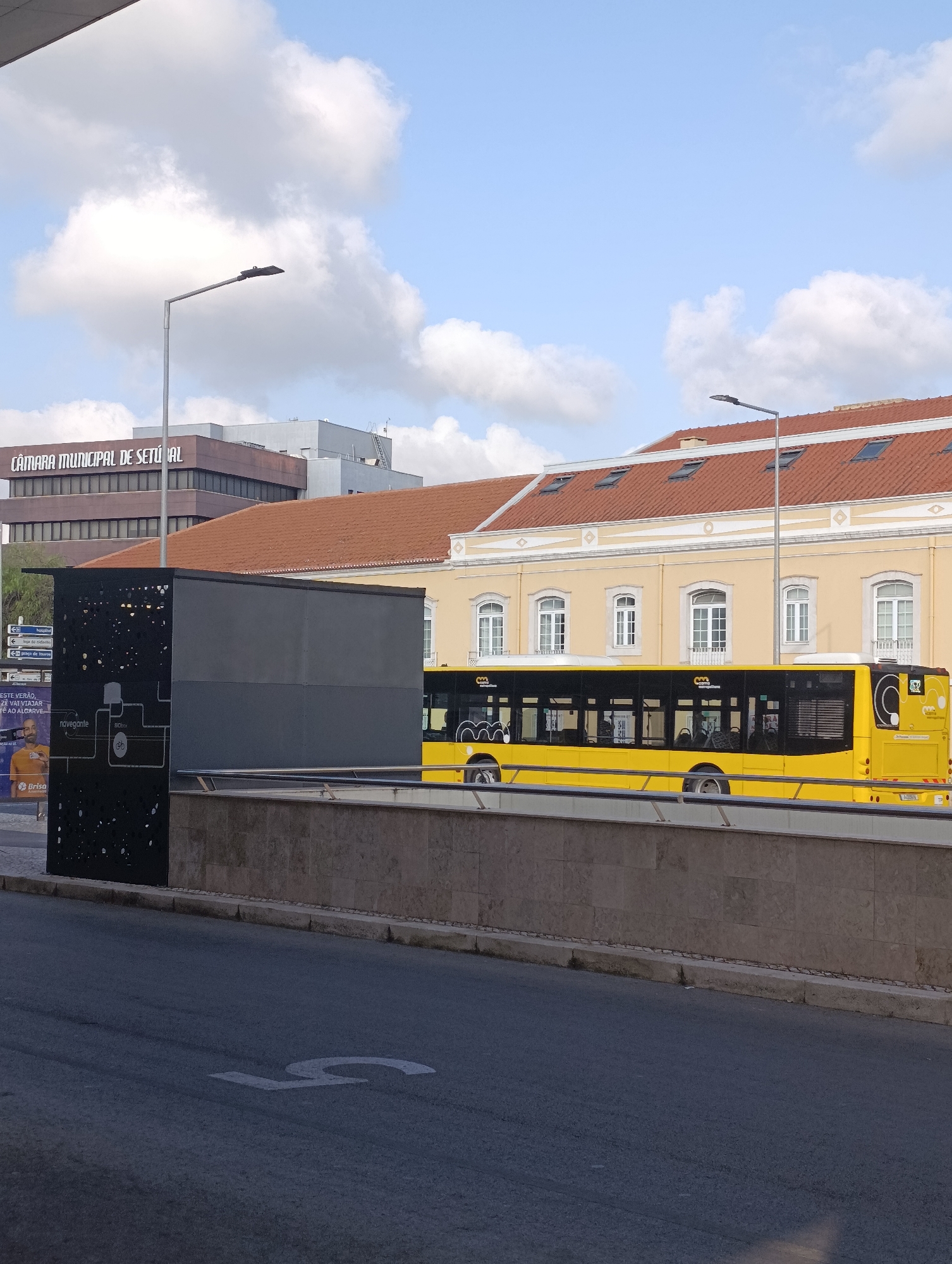 This is a full-shot, eye-level, outdoor photograph of a street scene in Setúbal, Portugal, on a partly cloudy day. The primary subjects are a modern yellow bus and a building with a terracotta roof.
In the foreground, a dark asphalt road features a large white "5" painted on its surface. A light-colored concrete wall runs along the edge of the road, with a metal railing on top. To the left of the wall, a tall, dark gray, rectangular structure stands, adorned with a white, schematic-like graphic. Part of an advertisement with a man's face and text in Portuguese is visible behind this structure.
A bright yellow bus, with black-tinted windows and white detailing, is the most prominent object. It is positioned on a road beyond the wall, and its side is visible. The bus has several visible advertisements or logos on its exterior.
Behind the bus, a building with a prominent terracotta-tiled roof and pale yellow walls is visible. The building has several evenly spaced windows with white frames and decorative elements above them. A taller, darker building with the words "CÂMARA MUNICIPAL DE SETÚBAL" (Municipal Chamber of Setúbal) in white lettering is located behind and to the left of the yellow-roofed building. Another gray, modern building is visible behind the Municipal Chamber building.
A streetlamp with a modern, flat design is positioned in the upper left quadrant of the image, extending over the scene. The sky is blue with scattered white clouds. No people are clearly visible.