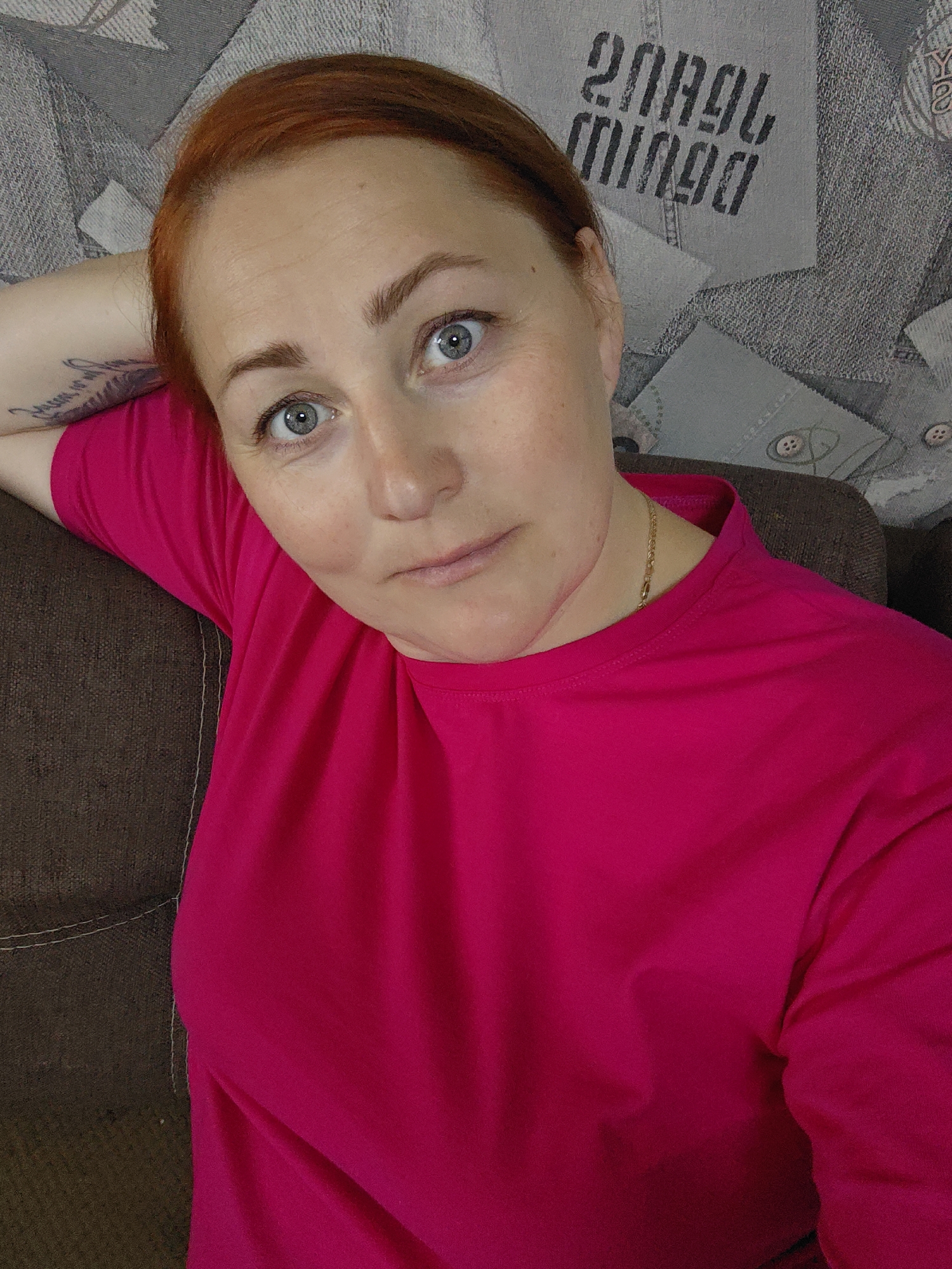 A medium-shot, selfie-style photograph depicts a woman with fair skin, light blue eyes, and reddish-auburn hair. Her hair is pulled back, revealing her forehead. She is wearing a bright pink t-shirt and a thin gold chain around her neck. Her left arm, adorned with a dark tattoo, is draped over the back of what appears to be a brown sofa or chair. The background consists of a wall covered in a patterned wallpaper depicting buttons and text in the style of denim clothing. The photograph was likely taken in an interior setting. There is no indication that the location is Марковский, Russia.