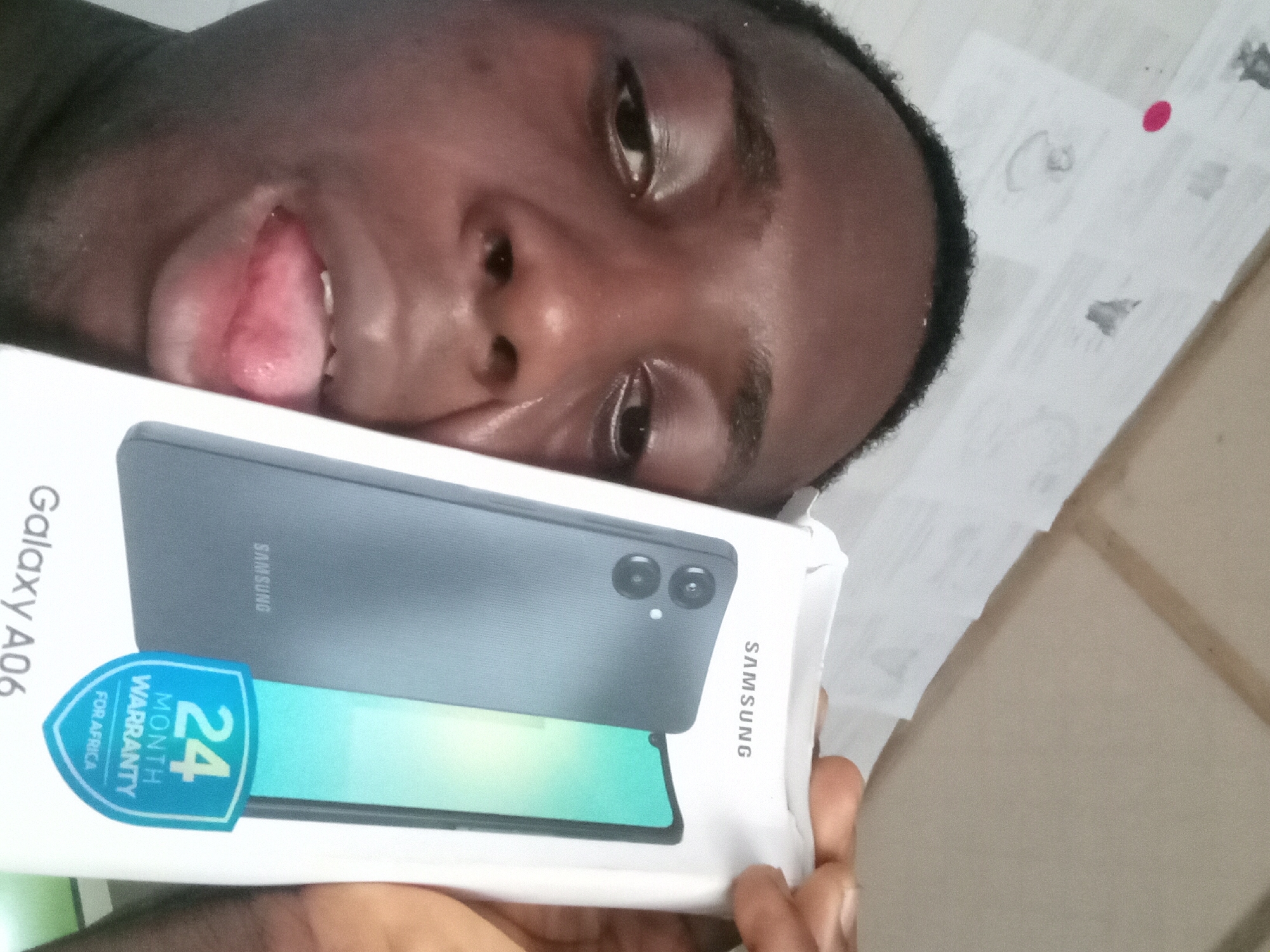A person is holding a Samsung Galaxy A06 phone box in an indoor setting. The person has dark skin, is sticking their tongue out, and has a close-up view of their face. The phone box has a picture of the phone on it and a blue sticker indicating "24 MONTH WARRANTY FOR AFRICA." The background is a white surface with some markings, potentially tile flooring. The location is not specified in the image, and information about Akwete, Nigeria is not apparent.