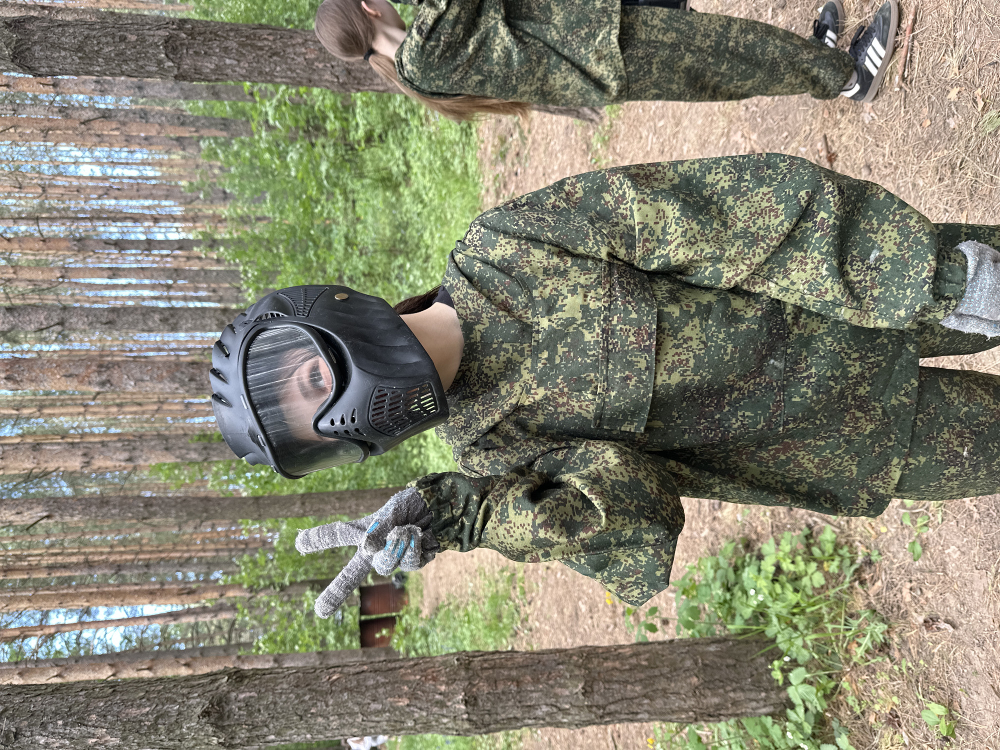 A paintball player in camouflage clothing and a black mask stands in a wooded area, making a peace sign with their gloved hand. Another person in similar camouflage clothing is partially visible in the background. The location appears to be a forest or park setting, possibly on the outskirts of Votkinsk, Russia, indicated by the natural environment and the potential for recreational activities like paintball. The ground is covered with dirt, fallen leaves, and some vegetation. The trees are tall and thin, typical of a coniferous forest. There are no buildings or urban infrastructure visible in the frame.