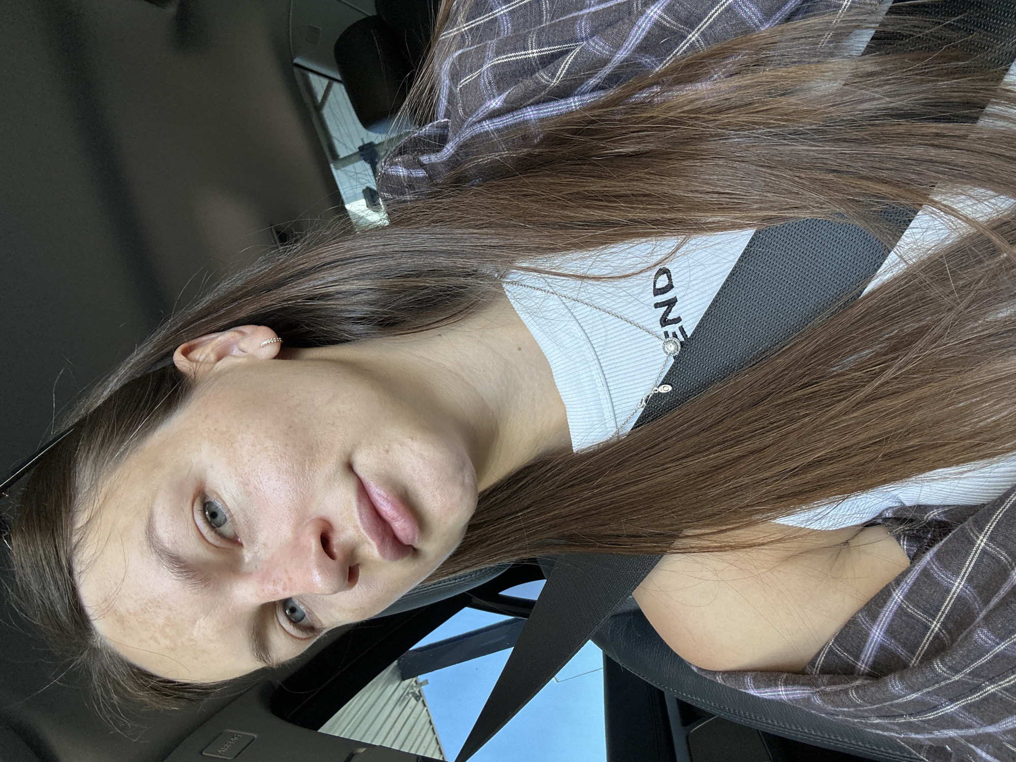 A young woman with long, brown hair is seated in the passenger seat of a car. She is wearing a white tank top with the word "END" printed on it and a plaid shirt draped over her shoulders. A seatbelt is fastened across her chest. The interior of the car is visible, as is a glimpse of the sky and a building through the window. The image is a selfie taken from a slightly high angle.