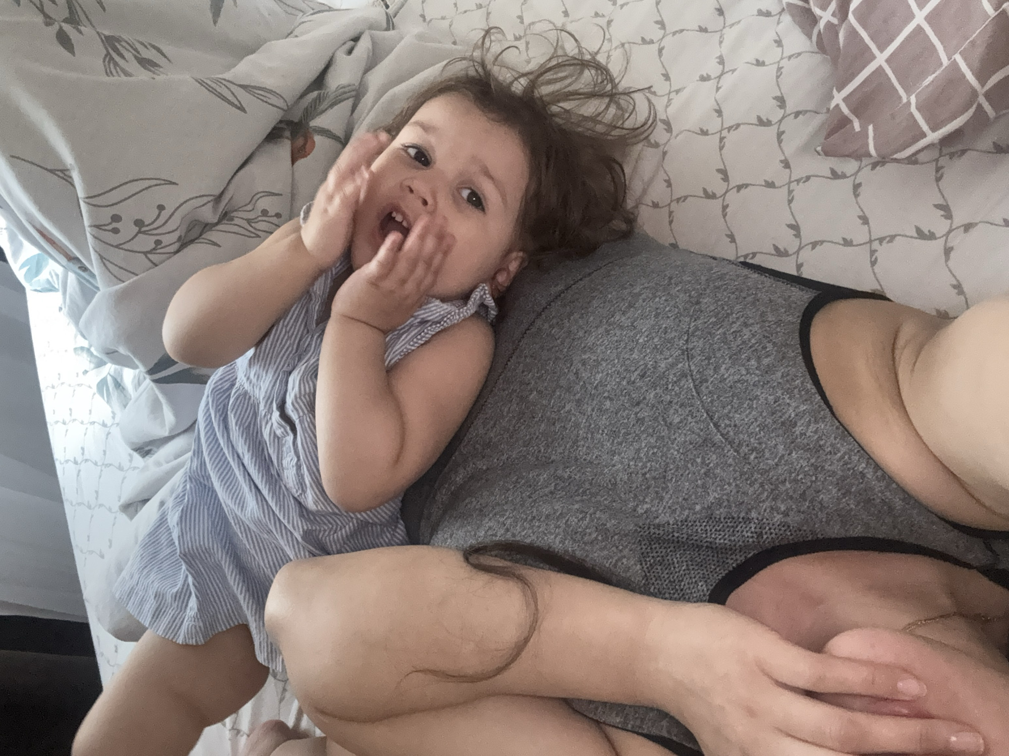A high-angle shot shows a young child with brown hair, wearing a blue and white striped romper, lying on a bed with a person wearing a gray shirt. The child has both hands raised towards their face, with a visible open mouth. The bed has a light-colored patterned sheet and a pillow with a geometric design. The location cannot be determined with the provided information.
