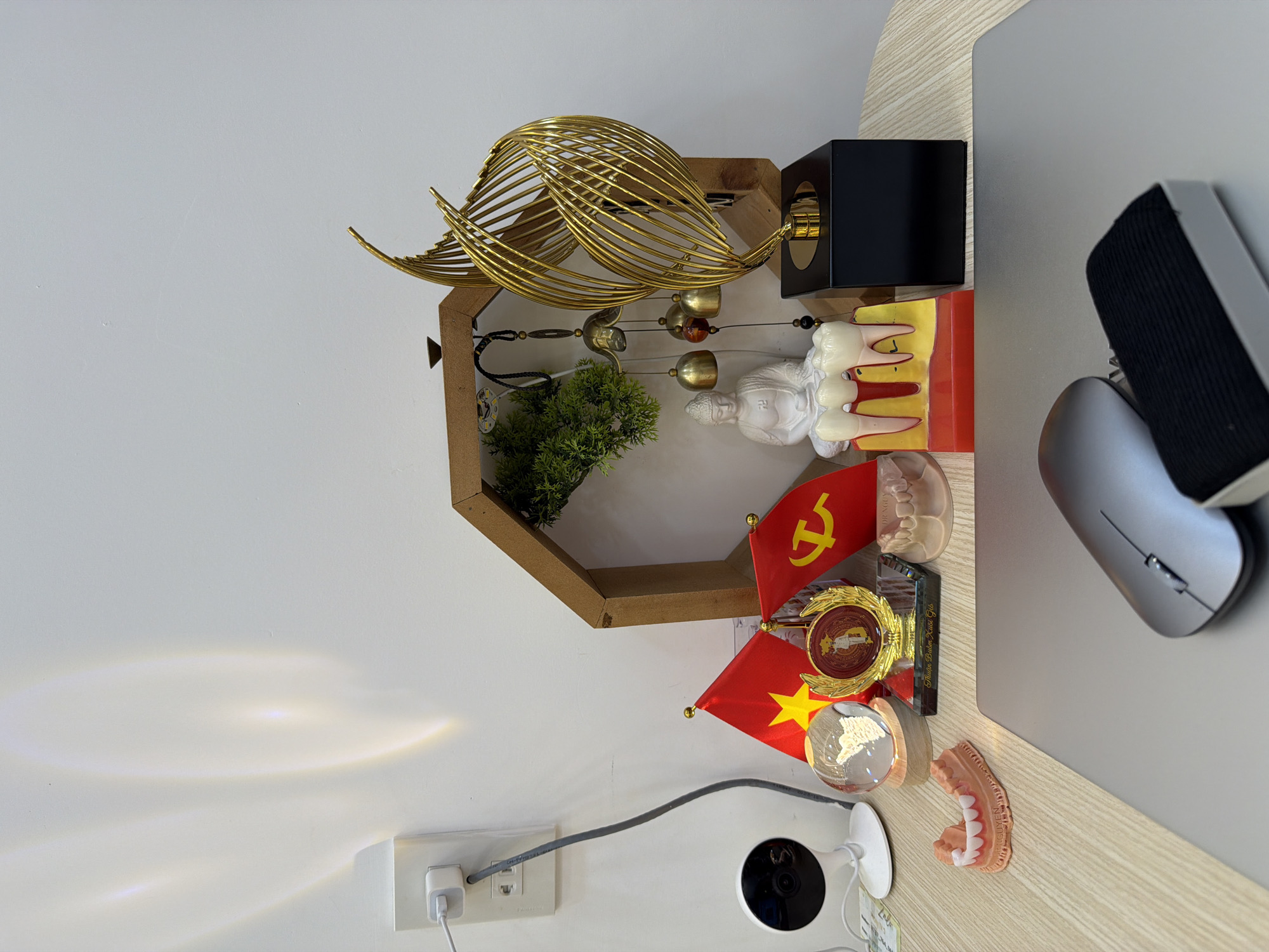 The image shows a shelf with various items displayed. On the shelf, there is a golden wire sculpture, a small Buddha statue, a miniature artificial plant, and two Vietnamese flags. Beneath the shelf, there are more decorative items including a crystal sphere, a trophy, and a model of teeth. To the right of the shelf, a computer mouse and a charging cable are visible, connected to a white power outlet. The scene appears to be an indoor setting, possibly a desk or a display area, with a light source casting a colorful glare on the wall in the background. The overall impression is of a collection of personal items or mementos.