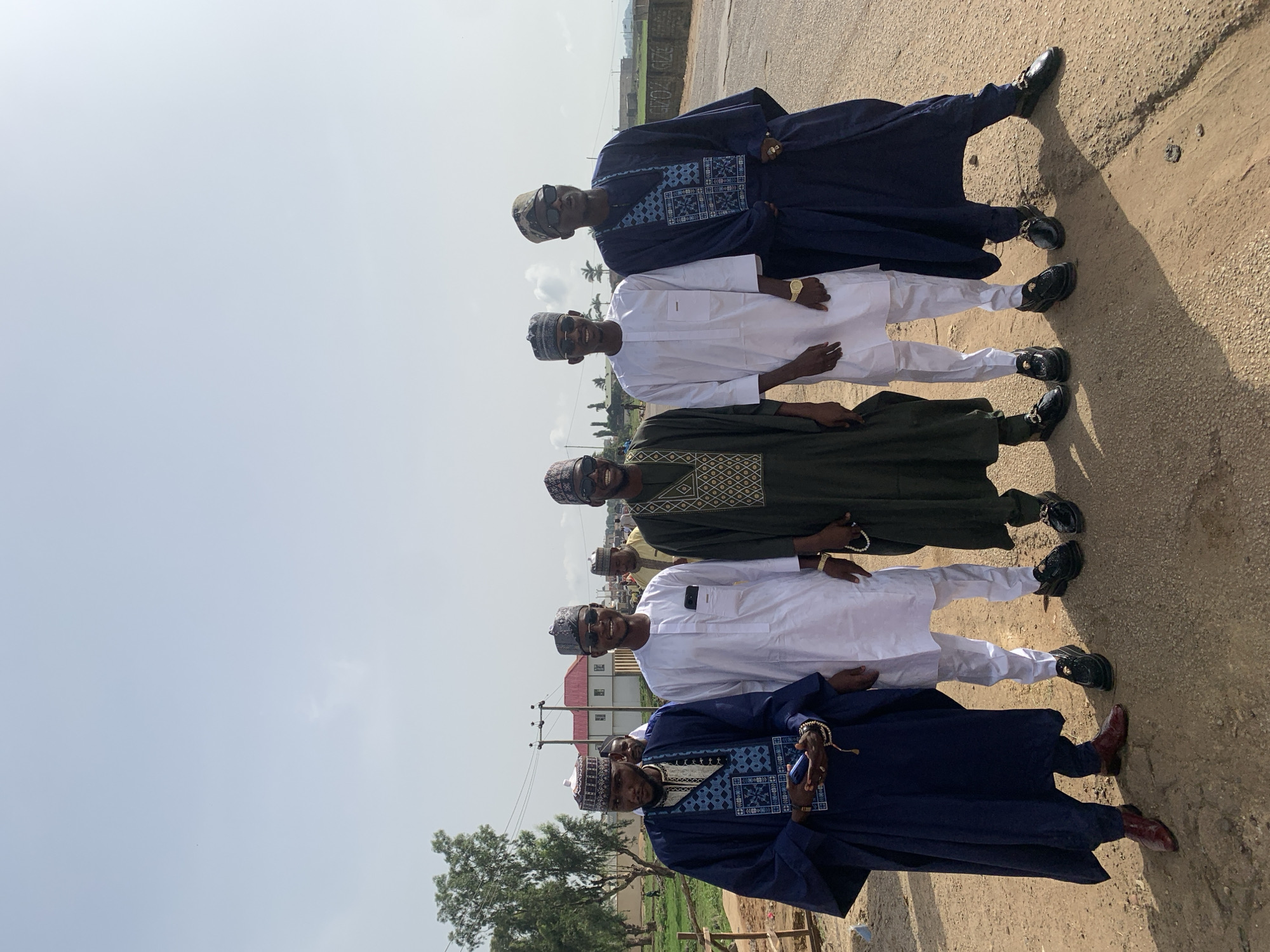 Five men are standing shoulder-to-shoulder on a paved surface in Naraguta Hausawa, Nigeria. Each man is wearing a traditional Muslim outfit. The first and last men are wearing dark blue robes with intricate patterns on the chest and sleeves and coordinating embroidered caps. The second and fourth men are wearing white robes and pants, and matching caps. The man in the middle is wearing a dark green robe with an embroidered chest and a matching cap. All the men are wearing sunglasses. The surface they are standing on appears to be a road or path. In the background, there are buildings, trees, and power lines. The sky is clear.