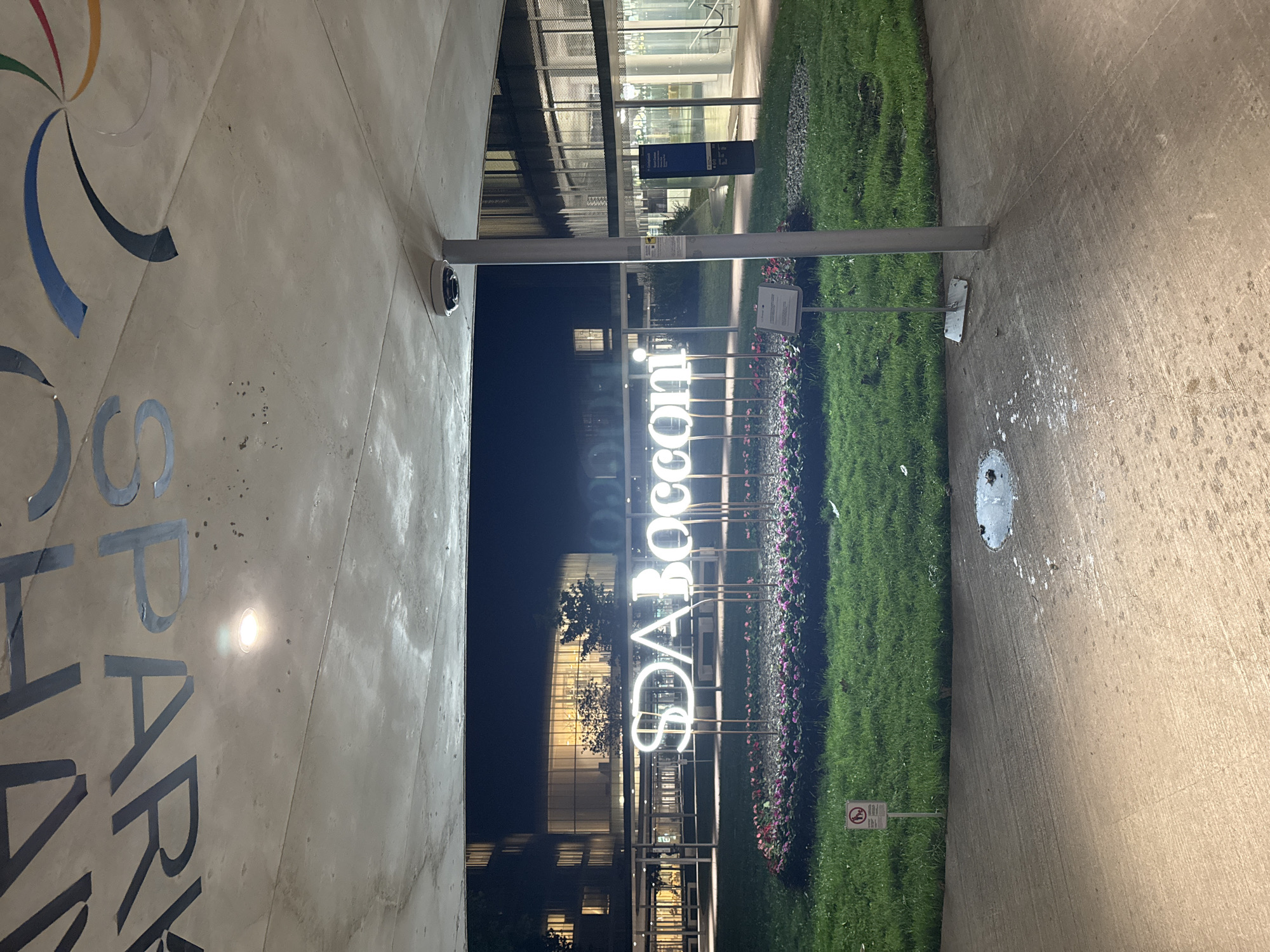 This nighttime shot depicts the campus of Bocconi University in Milan, Italy. The "SDA Bocconi" signage is prominently featured, illuminated against the backdrop of the campus buildings. To the left, partial lettering suggests branding related to the university. A strip of green lawn runs vertically between the signage and a paved area. The scene is lit artificially, with streetlights and building lights contributing to the overall illumination.