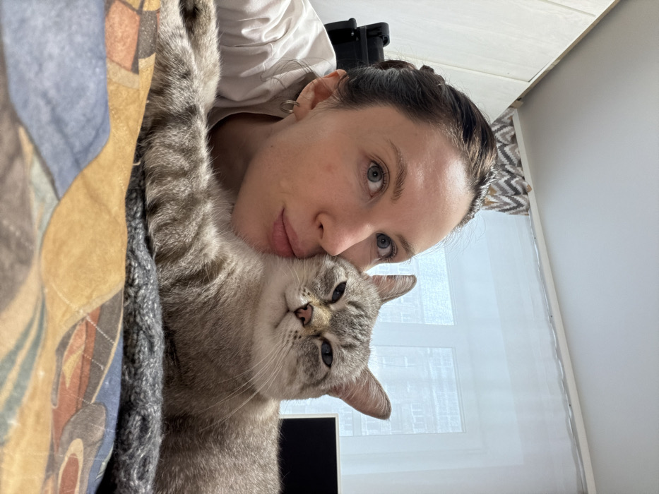This indoor photograph, taken in Perm, Russia, depicts a female and a cat in close contact. A Caucasian female, with dark hair pulled back and light eyes, wearing a light-colored shirt and small hoop earrings, leans into the frame from the left, presenting her profile. Her lips are puckered against the cheek of a light grey tabby cat with darker stripes. The cat is lying on a dark grey textured blanket, its front paws extended, with partially closed eyes. Both subjects are positioned on a patterned comforter displaying geometric designs in shades of blue, orange, brown, and grey. In the background, a large window covered by sheer white curtains reveals the exterior of an urban residential building. Above the window, a zigzag patterned valance in shades of brown, white, and grey is visible. To the left, a light-colored wall or cabinet and a dark, indistinct object are partially visible. To the right, a portion of a dark computer monitor is present.