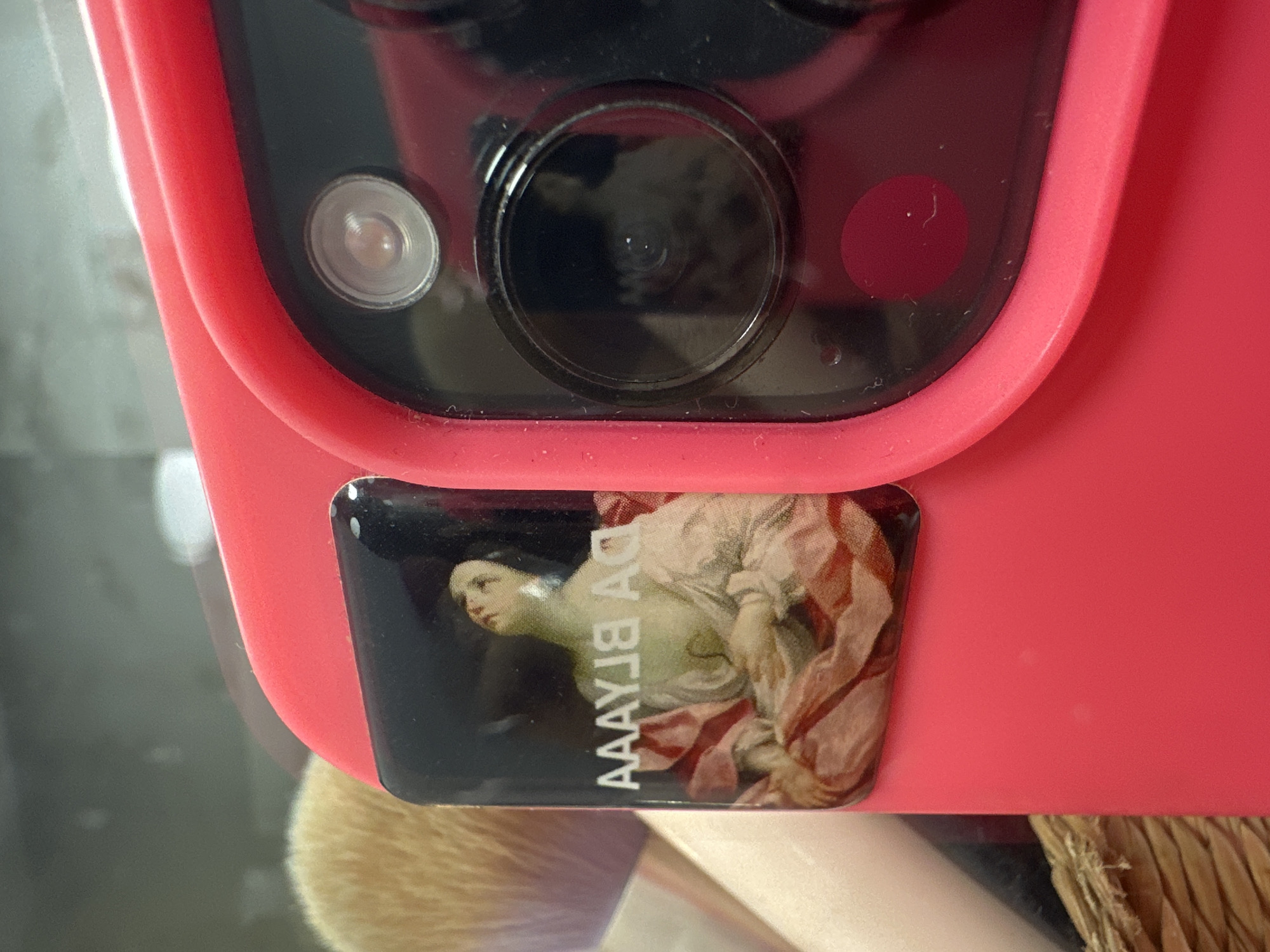 The image shows a close-up of a red phone case, likely an iPhone due to the distinctive camera module with three lenses and a flash. A small, rectangular sticker is affixed to the case, depicting a classical painting of a woman and the text "AAAЯJA DA BLAAYA". The background is blurry, with a makeup brush partially visible and some kind of woven basket texture. The scene is not identifiable as being located in Budva, Montenegro.