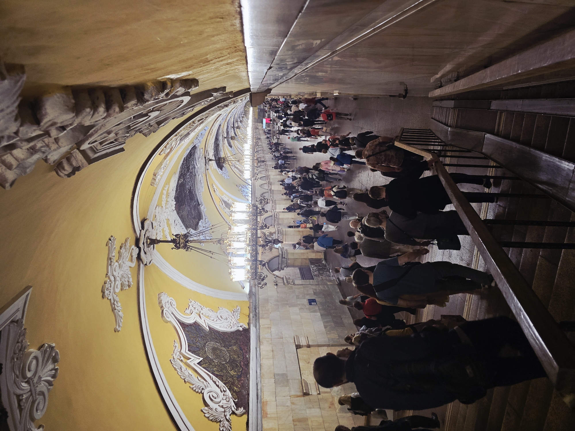The image depicts a crowded platform in a Moscow metro station, potentially the Kiyevskaya station, recognizable for its ornate, baroque architectural style. Passengers of diverse ages and attires fill the platform, suggesting peak hours. The yellow-painted walls are decorated with plasterwork and mosaics. Large chandeliers hang from the arched ceiling. An escalator leads down to another level.