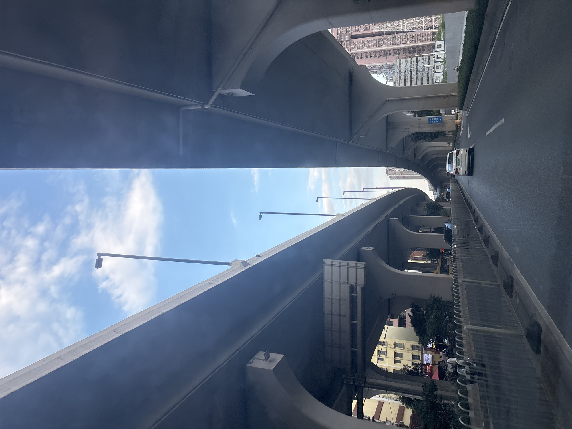 The image captures a view from within a vehicle driving on a multi-level highway interchange, likely in Baiyun District, China, given the architectural style and presence of vehicles. Overhead concrete overpasses curve into the distance, supported by massive concrete pillars. The sky is blue with scattered white clouds, and a few streetlights are visible along the upper level of the highway.
Below the overpasses, a road surface is visible with lane markings. A white car is driving ahead in the right lane. To the left of the road, there is a metal fence and some greenery. In the background, apartment buildings with many windows are visible on the right, suggesting an urban environment. There are also some electrical towers or structures in the distance. The overall impression is one of modern urban infrastructure.