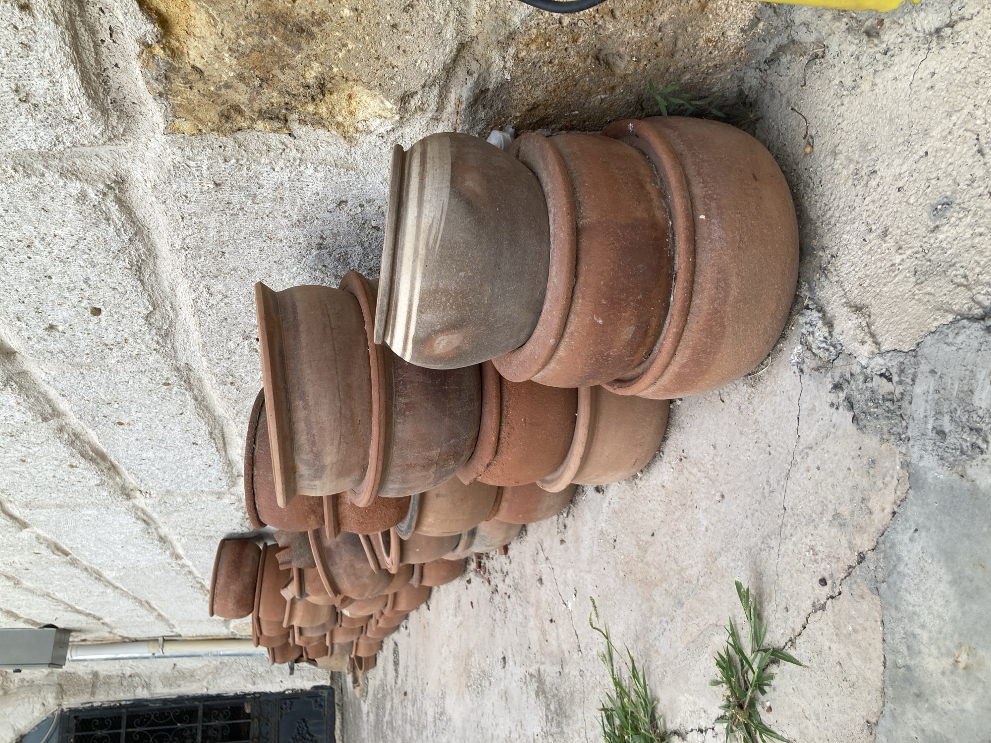 A stack of earthenware pots is displayed outside on a textured concrete surface in front of a stone wall. The pots vary in size and shape, with the majority being reddish-brown in color, though some exhibit a lighter, grayish hue. They are arranged in an overlapping fashion, creating a visually dense composition. In the background, a portion of a building is visible, featuring a dark metal gate and a light-colored stone facade. Green grass grows sporadically through cracks in the concrete in the foreground and to the side of the pots. The scene suggests an outdoor market or shop in Avanos İlçe Merkezi, Turkey, known for its pottery. There are no people or animals present.