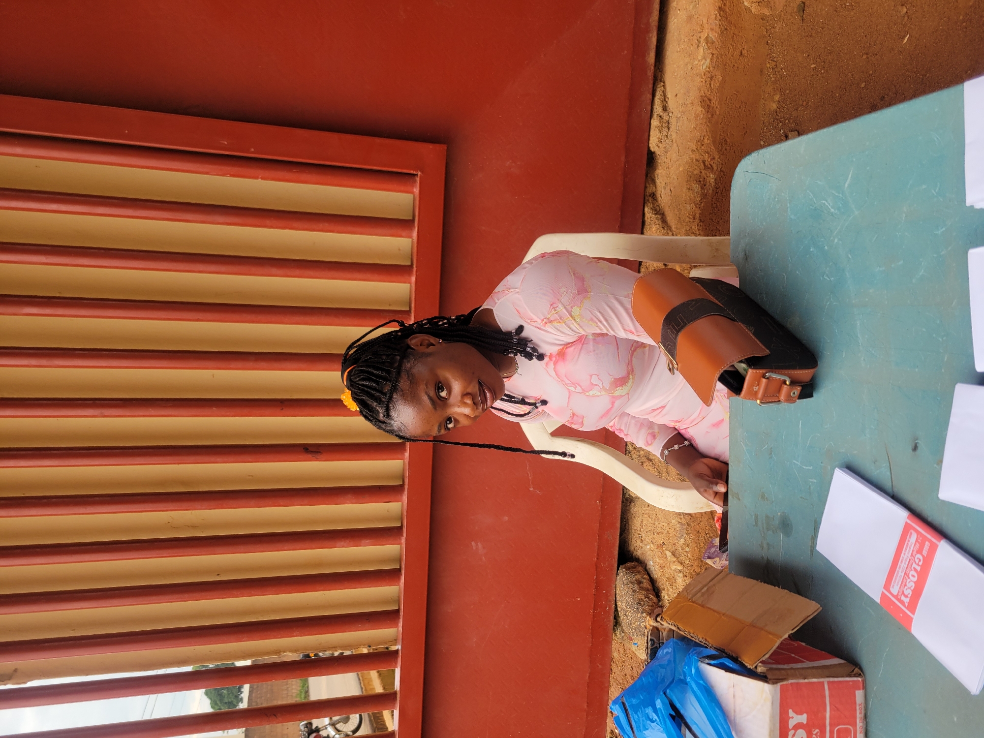 The scene is set outdoors in Gura Top, Nigeria, featuring an adult female seated at a table against a building or wall. The woman, of African ethnicity, wears her dark hair in braids, secured with a yellow accessory near the crown. She is dressed in a light-colored, long-sleeved garment with a subtle pattern and has a bracelet on her left wrist. She is seated in a light-colored plastic chair, looking directly forward.
In the foreground, a blue-green rectangular table holds several items. On the left side, a partially open cardboard box displays the partial text "SY" and "NO. IN BOX", with blue plastic material visible within or beside it. To the right of the box is a stack of white, rectangular paper or card stock banded with a red label that reads "GLOSSY". A brown and black shoulder bag with a visible buckle and strap rests further right on the table. A smaller stack of white papers is also visible on the far right of the table.
The background consists of a reddish-brown wall, above which is a section with vertical red bars set against a lighter, possibly tan, background. This arrangement gives the appearance of a barred window or gate. Through the bars on the left, a portion of an unpaved road or path is visible, along with foliage and the wheel of a motorcycle or bicycle. The ground beneath the table is unpaved earth, littered with small rocks and debris.