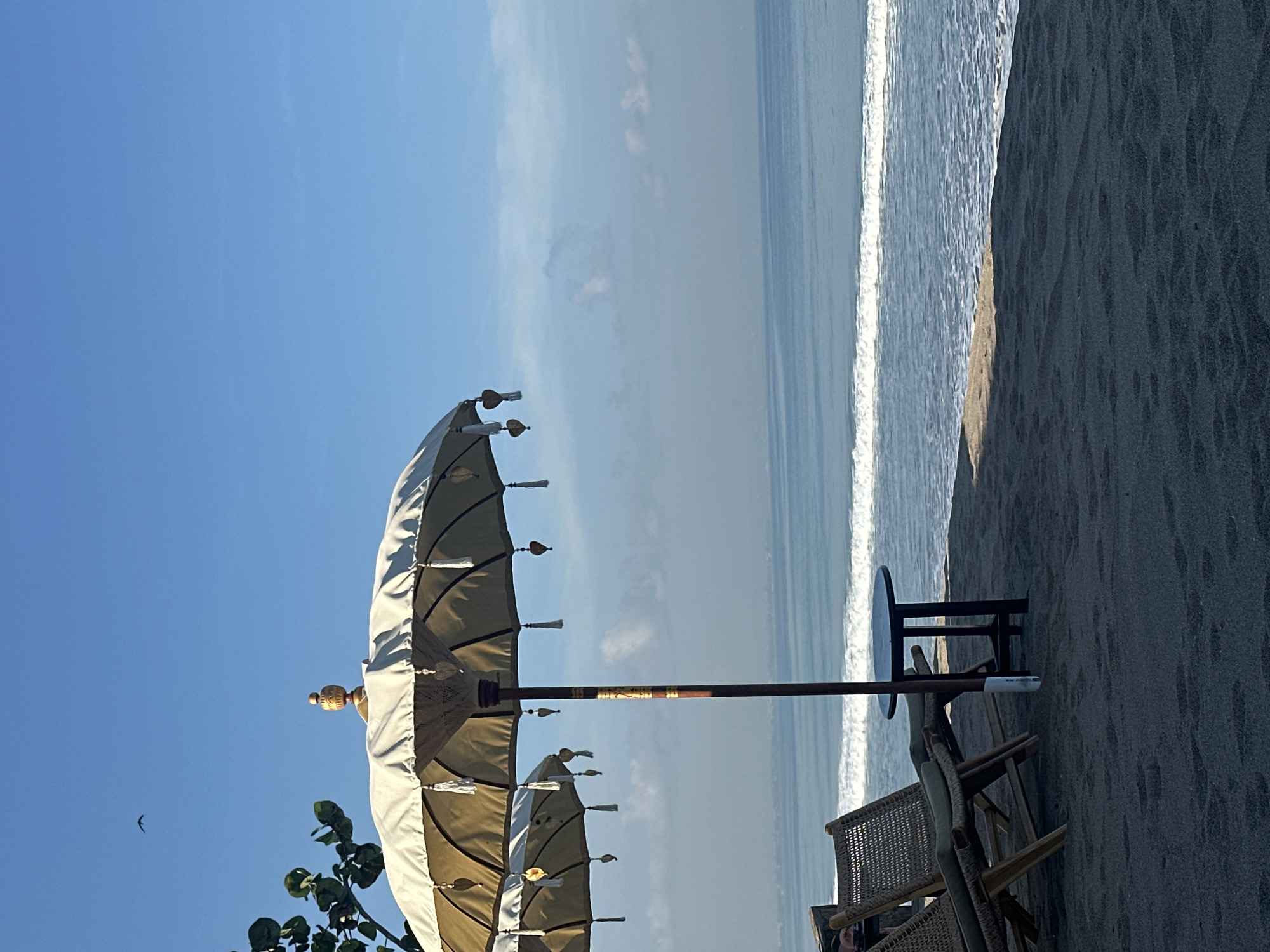 This image captures a serene beach scene in North Kuta, Indonesia, oriented in portrait mode, though presented rotated. The sky occupies the upper portion, transitioning to the ocean and a dark sand beach on the right.
In the foreground, slightly to the left, stands a tall, ornate beach umbrella. Its canopy is a light, perhaps off-white or beige, fabric, featuring decorative tassels and a golden, carved finial at its peak. The umbrella's central pole has intricate patterns or bands. Below it, some green foliage is visible. Another similar umbrella or a lower section of the main umbrella is partially seen beneath the primary one.
To the right of the umbrella, set on the dark sand, are a small, round, dark-colored table and two chairs. One chair, made of woven material and wood, is clearly visible, while the other is partially obscured behind it.
The mid-ground transitions into the ocean, characterized by gentle waves breaking on the shore, forming a distinct white line of foam along the water's edge. The ocean appears calm and extends into the distance under a bright sky. The far right of the image shows the dark, granular texture of the sand.
The sky is a clear, bright blue with a few faint, wispy clouds scattered across it. A small bird is silhouetted against the sky in the upper left corner, mid-flight. The sunlight appears bright, casting subtle shadows from the chairs and table onto the sand, suggesting either morning or late afternoon. The overall atmosphere is peaceful and inviting, typical of a relaxing beach setting. There is no visible text in the image.