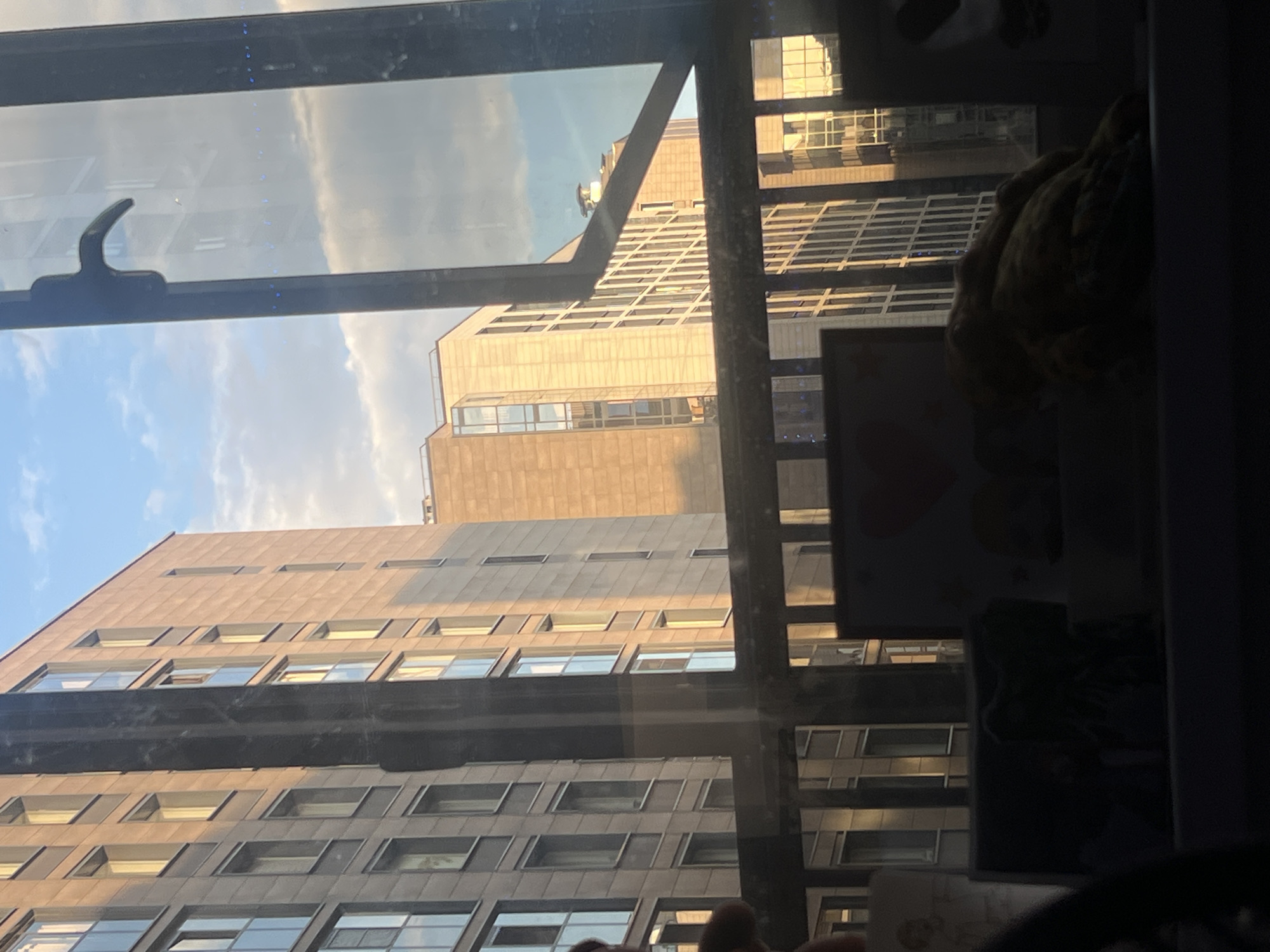 The image displays an exterior view of a city from a window in Saint Petersburg, Russia, captured during the late afternoon or early evening, characterized by the warm, golden light of the "golden hour." The scene shows two prominent multi-story buildings under a clear, light blue sky with scattered white clouds.
In the foreground, the dark frame of a window and a black window latch are visible on the left side. The window glass exhibits faint, small blue dots or lines arranged vertically, possibly reflections or details on the glass. Reflections of the sky and clouds are also visible on the pane. To the right, inside the room, indistinct, dark, and blurry objects are visible, including what appears to be a textured, soft item like a stuffed animal or blanket, resting near the window.
Outside, the main focus is on the architecture. A large, modern building occupies the left side of the frame, featuring numerous uniformly-sized rectangular windows set into a light brown or grey facade. This building is bathed in the warm, golden sunlight, creating a play of light and shadow on its surfaces. Further to the right and slightly behind, another tall building with a similar architectural style is visible, also illuminated by the low sun. The overall impression is one of urban tranquility during a beautiful sunset. No discernible text is visible in the image.