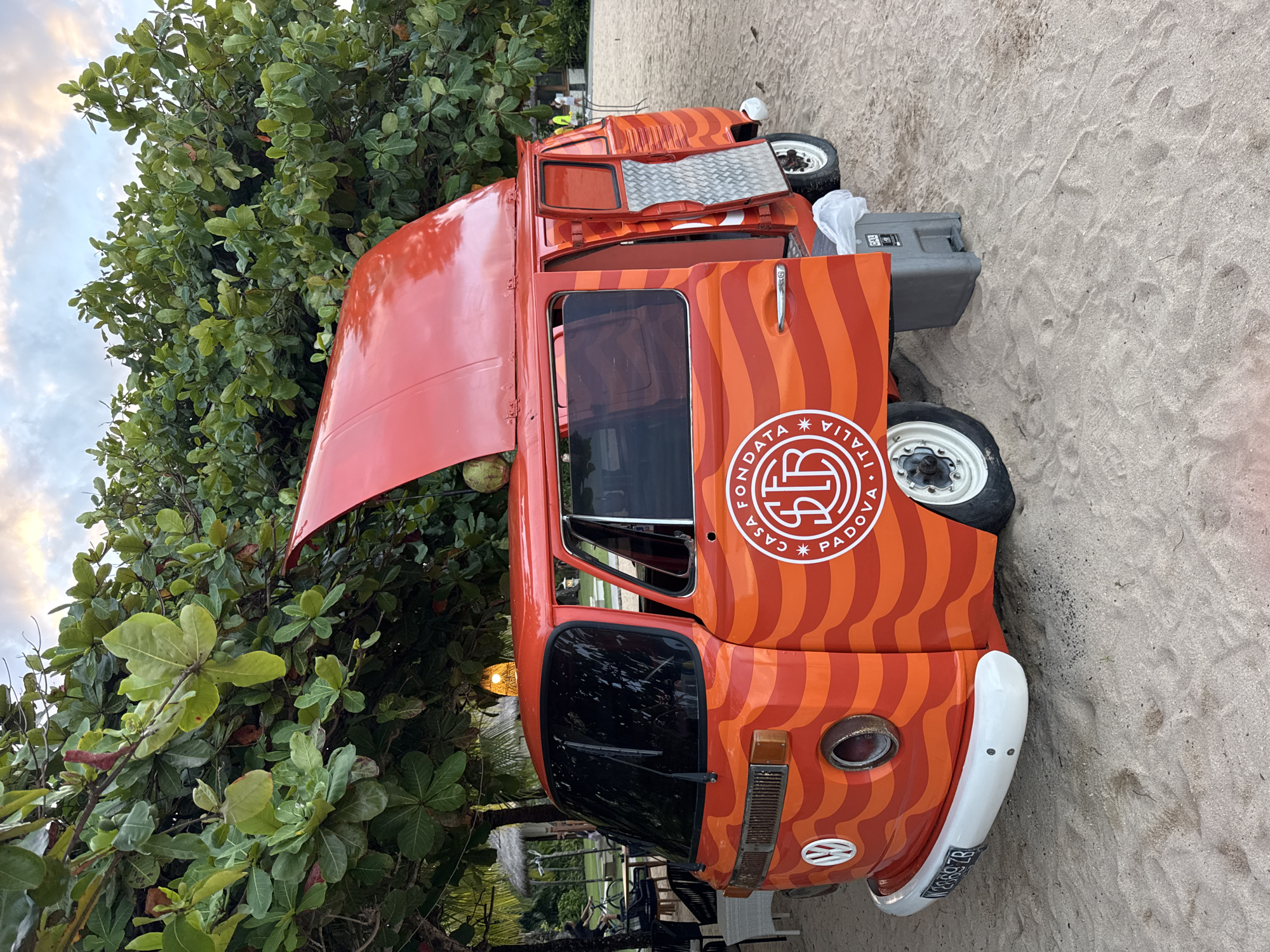 A medium shot of an orange and red vintage Volkswagen van parked on a sandy beach in Kuta Selatan, Indonesia. The van features a white circular logo with black text that reads "FONDATA CASA PADOVA ITALIA". The hood of the van is open, and a side door is ajar. Nearby, a green tree partially obscures the view, and a grey container rests on the sand near the vehicle. The beach is visible with light brown sand.