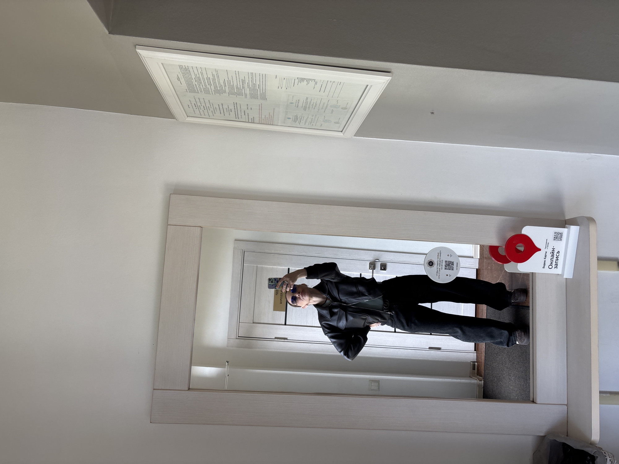 A woman is taking a selfie in front of a mirror. The mirror is hanging on a white wall. The woman is wearing a black jacket, black pants, and sunglasses. There is a picture on the wall above the mirror and a sign on the right side of the mirror with the words "Яндекс Карты" and "Онлайн-запись" in Russian.
