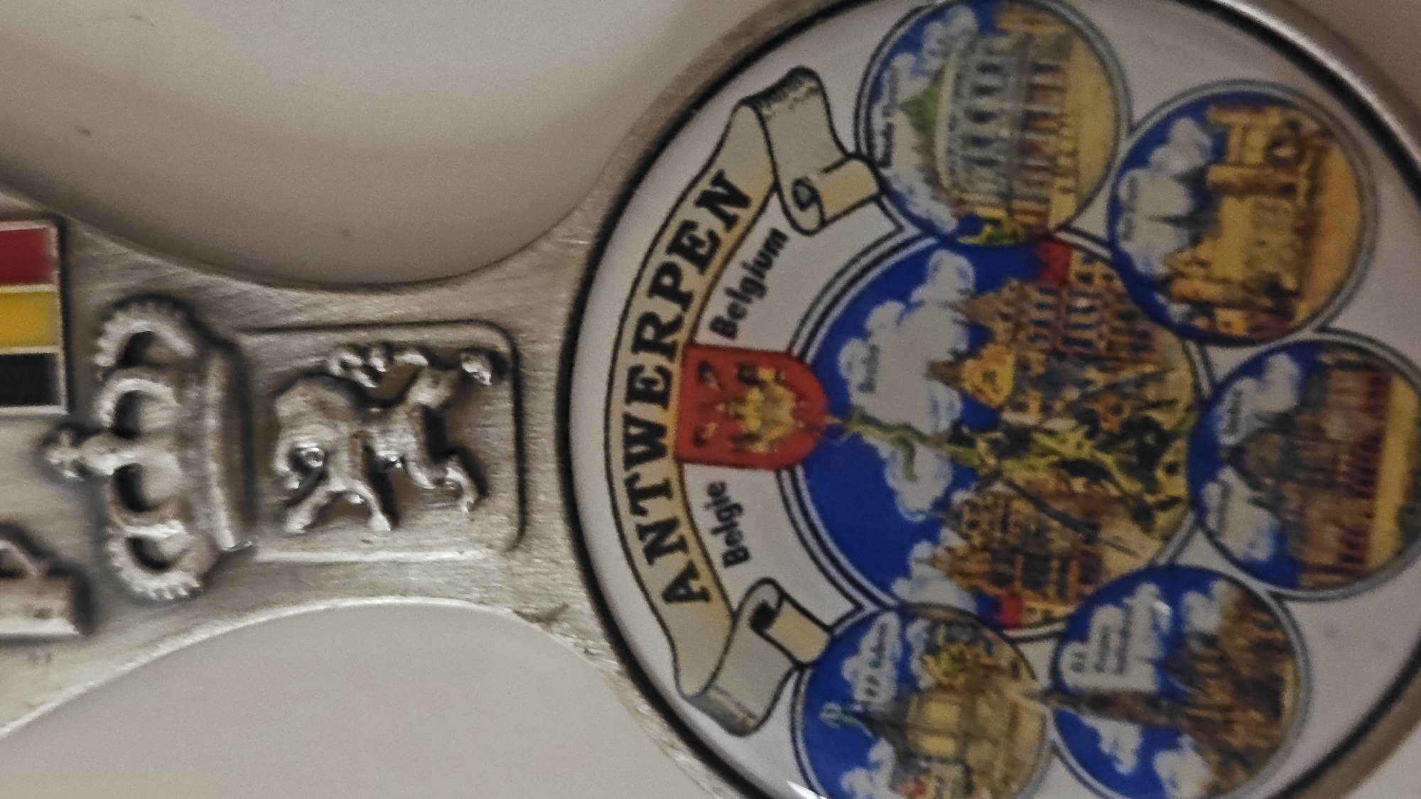 The image shows a souvenir spoon from Antwerp, Belgium. The spoon's head features a circular design with "Antwerpen" written on a banner above the city's coat of arms. Around the central depiction of Antwerp are smaller circular images of various buildings and landmarks. The spoon's handle displays the flag of Belgium, a crown, and another heraldic symbol. No part of the image is related to Sevastopol, Ukraine.