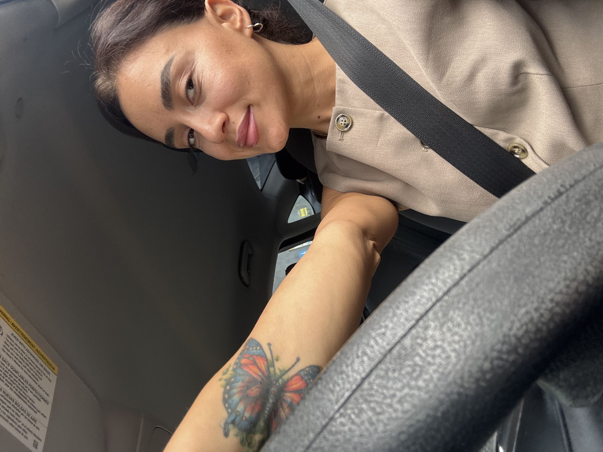 A woman with brown hair pulled back and a butterfly tattoo on her left arm is seated in the driver's seat of a car, looking towards the camera. She is wearing a tan jacket with a single button and a black seatbelt across her chest. The interior of the car is visible, including the steering wheel and the car's roof liner with warning labels. The scene is indoors, likely within a vehicle, with no discernible city or specific location indicated.