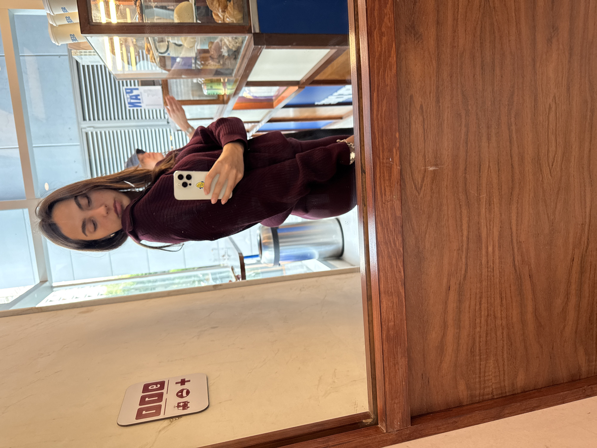 A young woman is reflected in a mirror. She has brown hair and is wearing a maroon sweater. She is holding a smartphone with a white case. She is taking a photo in what appears to be a cafe or bakery. The mirror is framed by a dark wood. In the background, there is a display case filled with pastries and other food items. Through the window, a view of the outside is visible. A sign with the name "Ojo de Agua" is seen on the countertop. This image was taken in Mexico City, Mexico.