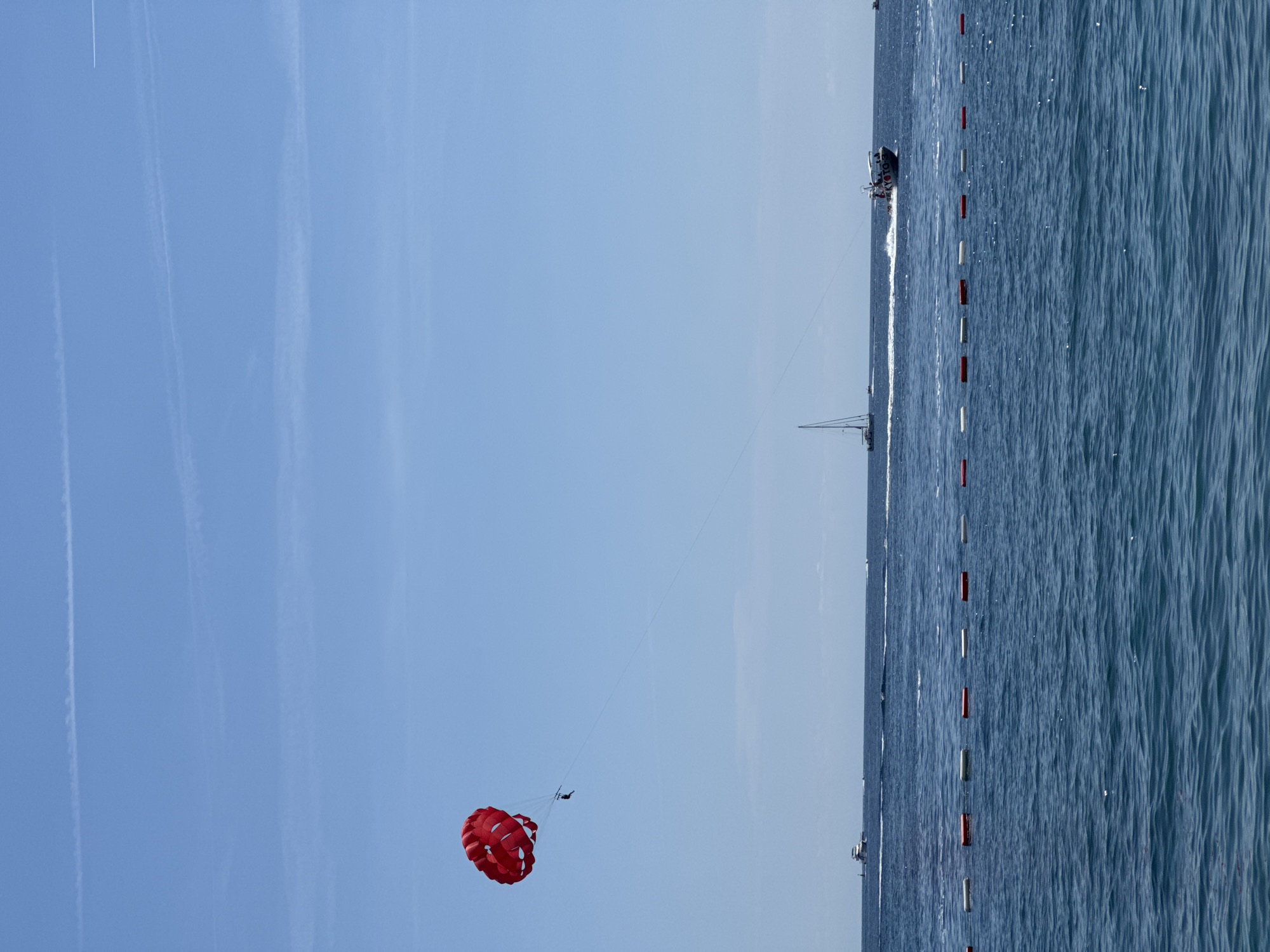 The image shows a parasailer soaring against a clear blue sky over the Adriatic Sea. A boat is visible towing the parasailer with a line. The boat is moving parallel to the coastline in Budva, Montenegro, as indicated by the presence of a line of buoys in the water.