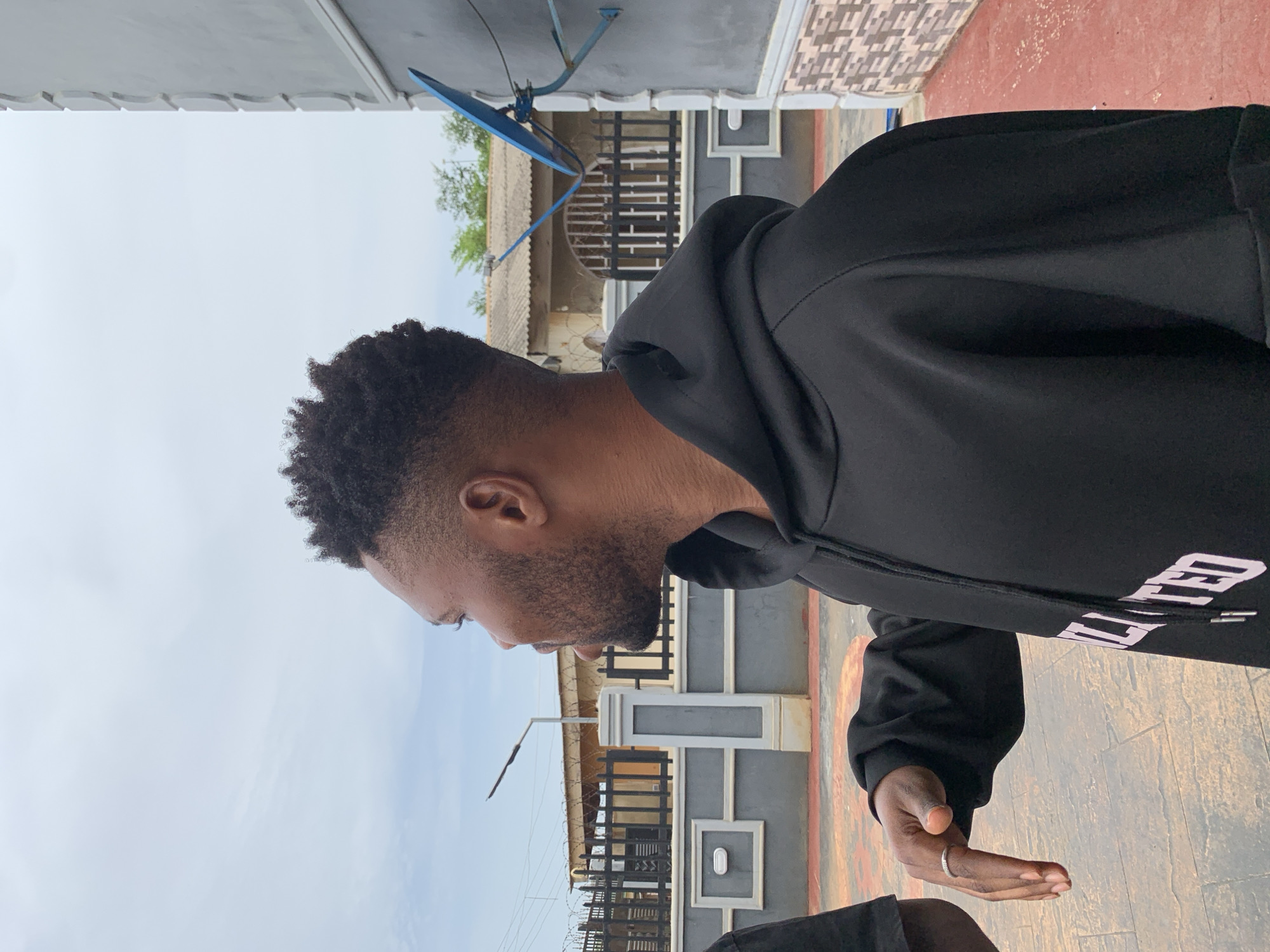 A man with dark skin, a trimmed beard, and short, dark, curly hair is captured in a profile shot, looking downwards. He is wearing a black hoodie with the word "ELEVATED" partially visible in white lettering on the side. A silver-colored ring is on the ring finger of his right hand, which is extended forward with an open palm.
The scene is an outdoor, daytime setting, likely a residential area in Ikorodu, Nigeria, under an overcast sky. In the background, a blue satellite dish is mounted high on a building. Below it, other structures include a building with a corrugated roof, and a modern grey and white fence or wall topped with a black gate and barbed wire. A street light pole is also visible. The ground in the foreground is paved with reddish-brown tiles, and a decorative grey and white wall with a light fixture runs along the side.